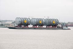 Barge carrying large gray cylindrical tanks with yellow accents on a body of water, likely a river.