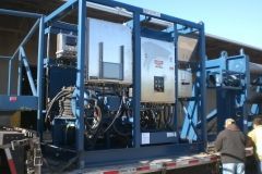 A large industrial machine in a blue metal frame on a flatbed trailer, with people nearby.