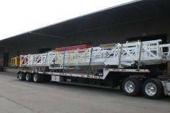 Large white metal structures loaded on a semi-truck trailer. Industrial setting, under a loading dock.