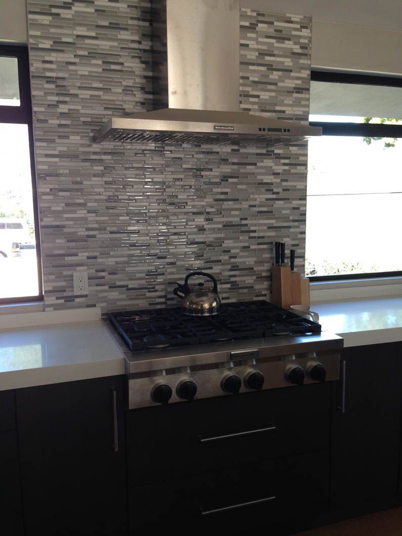 A stainless steel gas stove with a kettle sits beneath a vent hood against a gray, textured tile backsplash in a kitchen.