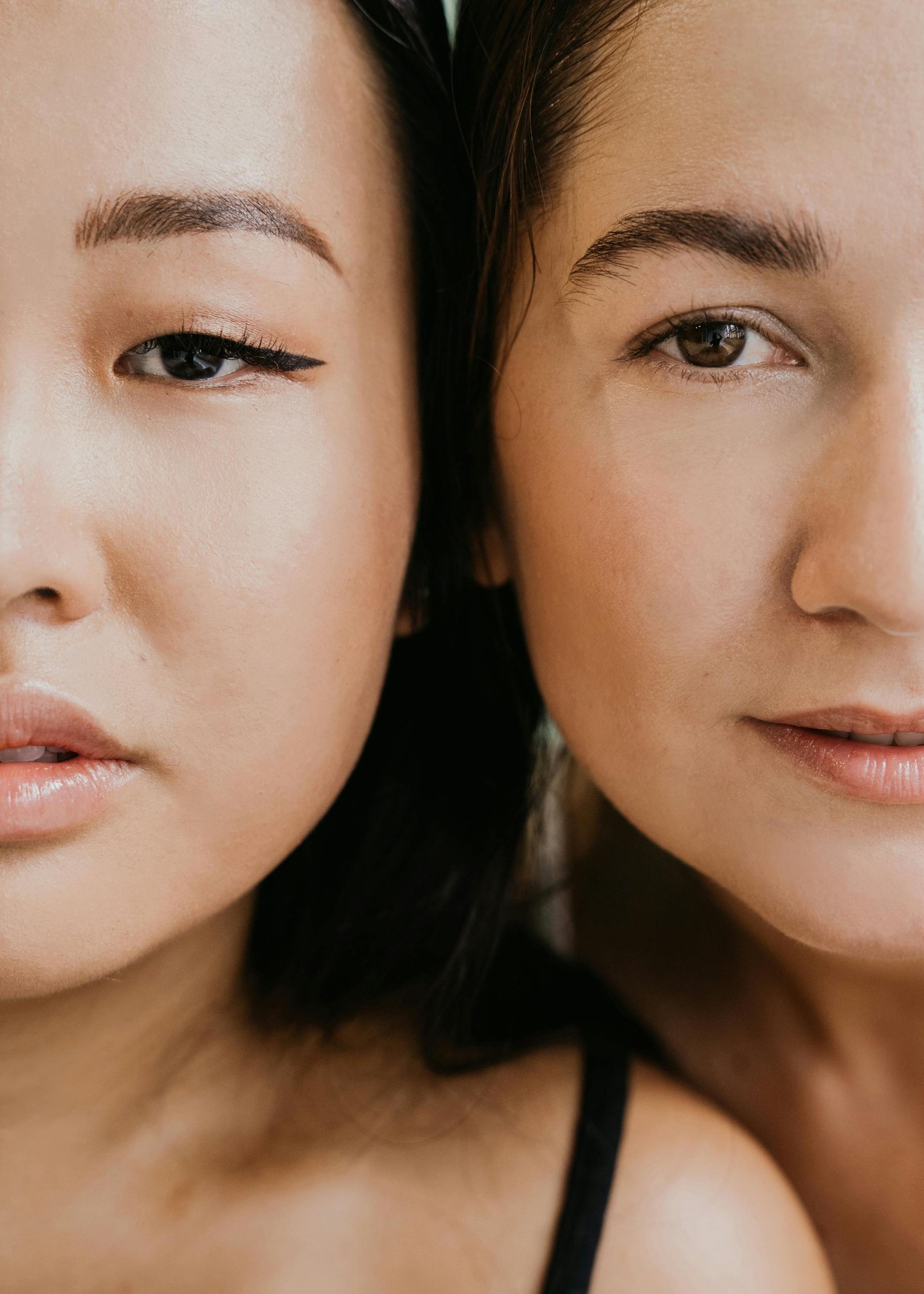 Two women, faces close together, looking at the viewer. One has eyeliner, the other, a neutral look.