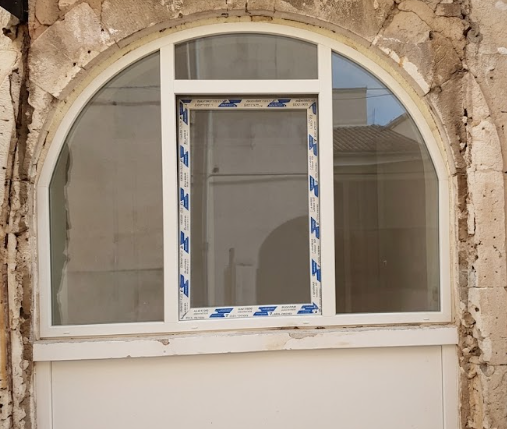 Finestra ad arco bianca incastonata in un muro di pietra consumato, con un vetro centrale ricoperto da nastro adesivo blu.