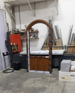 Officina con un grande telaio di porta ad arco in legno su un banco da lavoro, attrezzi e materiali sullo sfondo.
