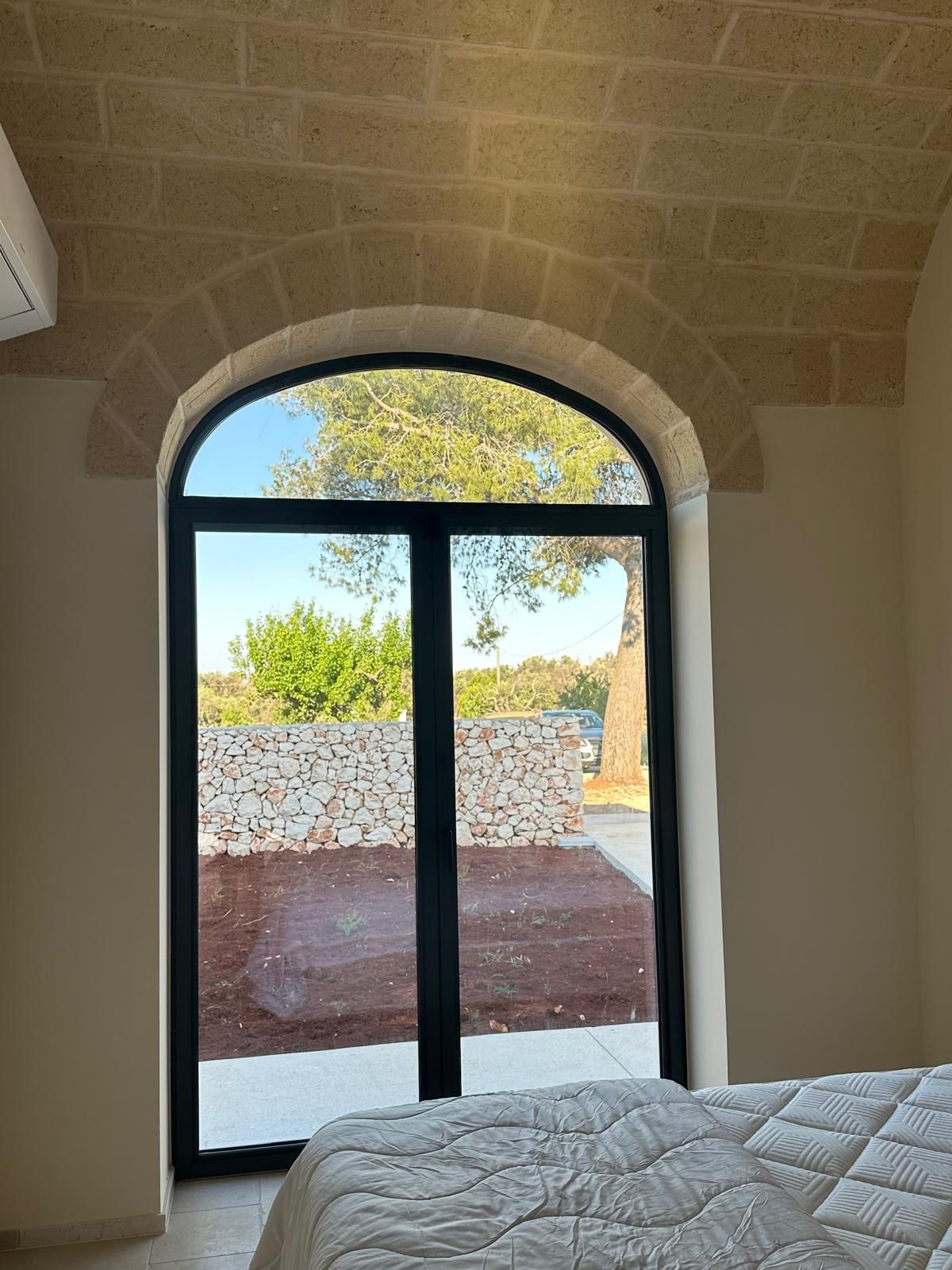 Camera da letto con porte finestre ad arco che si aprono su un patio soleggiato e vista sul muro in pietra.