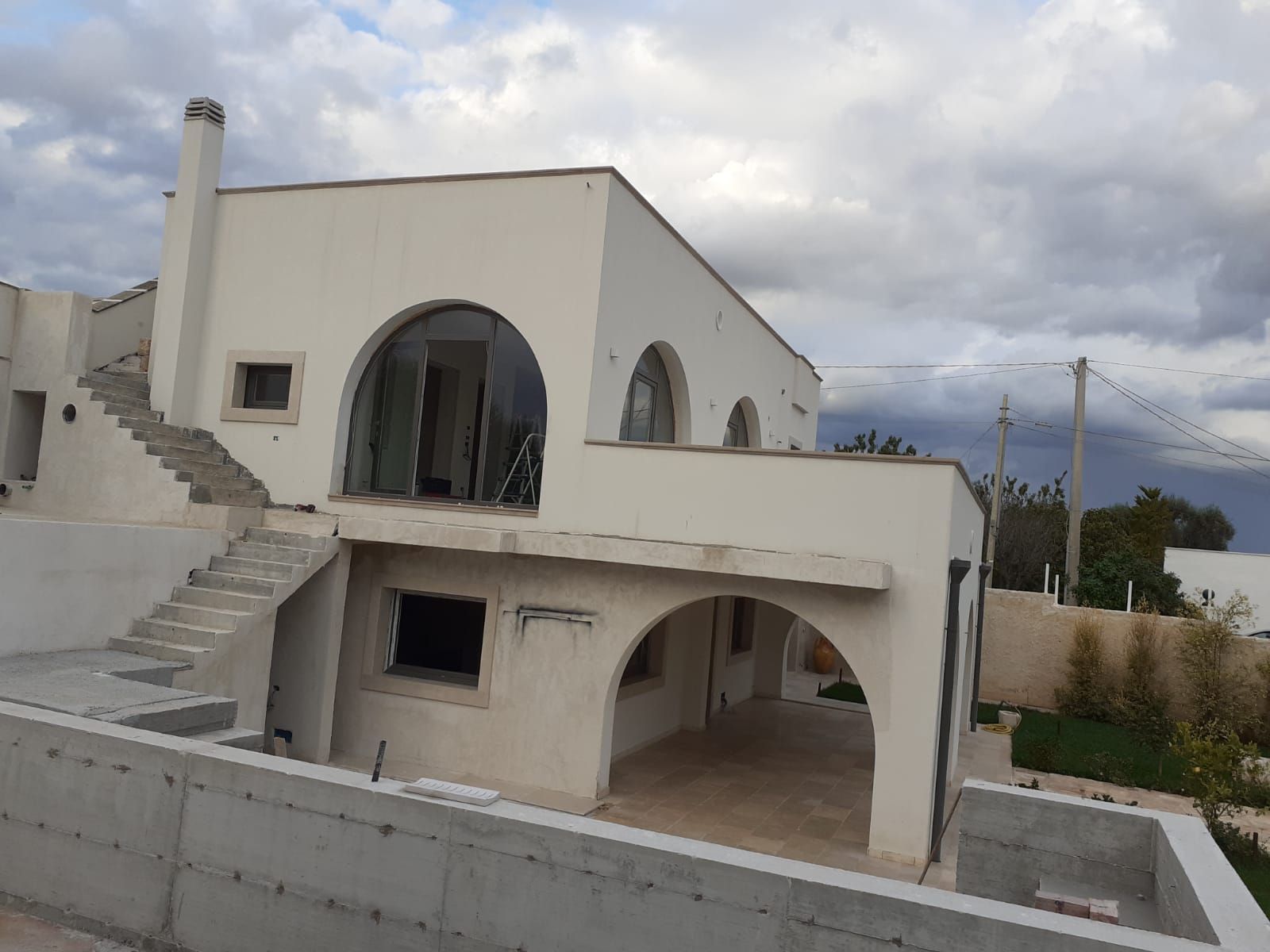 Casa bianca a due piani con finestre ad arco e scale, vista dall'esterno sotto un cielo nuvoloso.