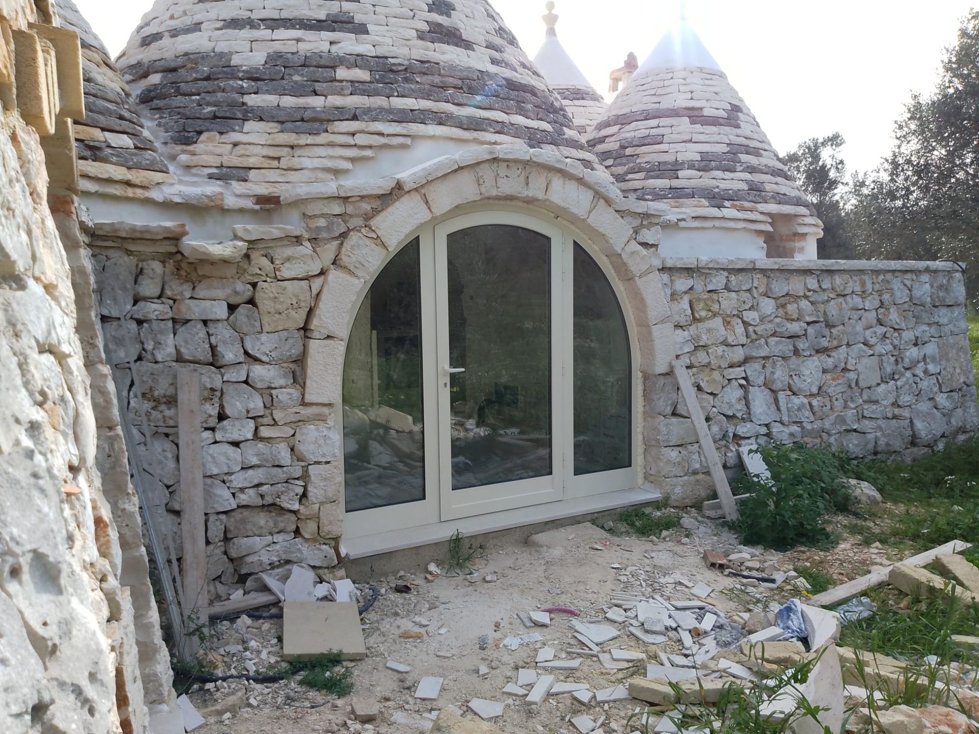 Casetta in pietra con porta ad arco bianca e muri in pietra grezza, circondata da detriti e vegetazione.