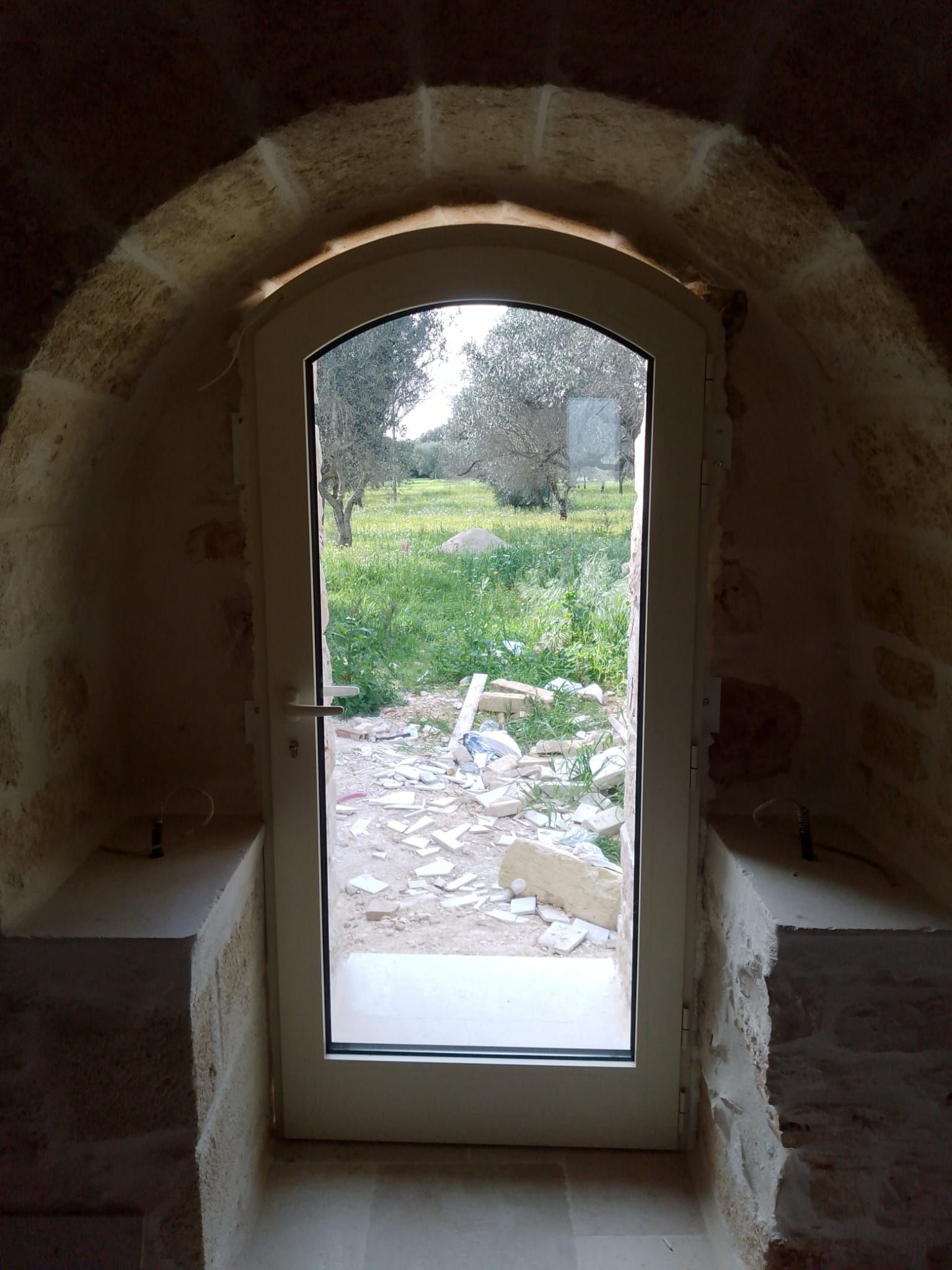 Finestra ad arco in un muro di pietra, che si affaccia su un cortile erboso con alberi e macerie.