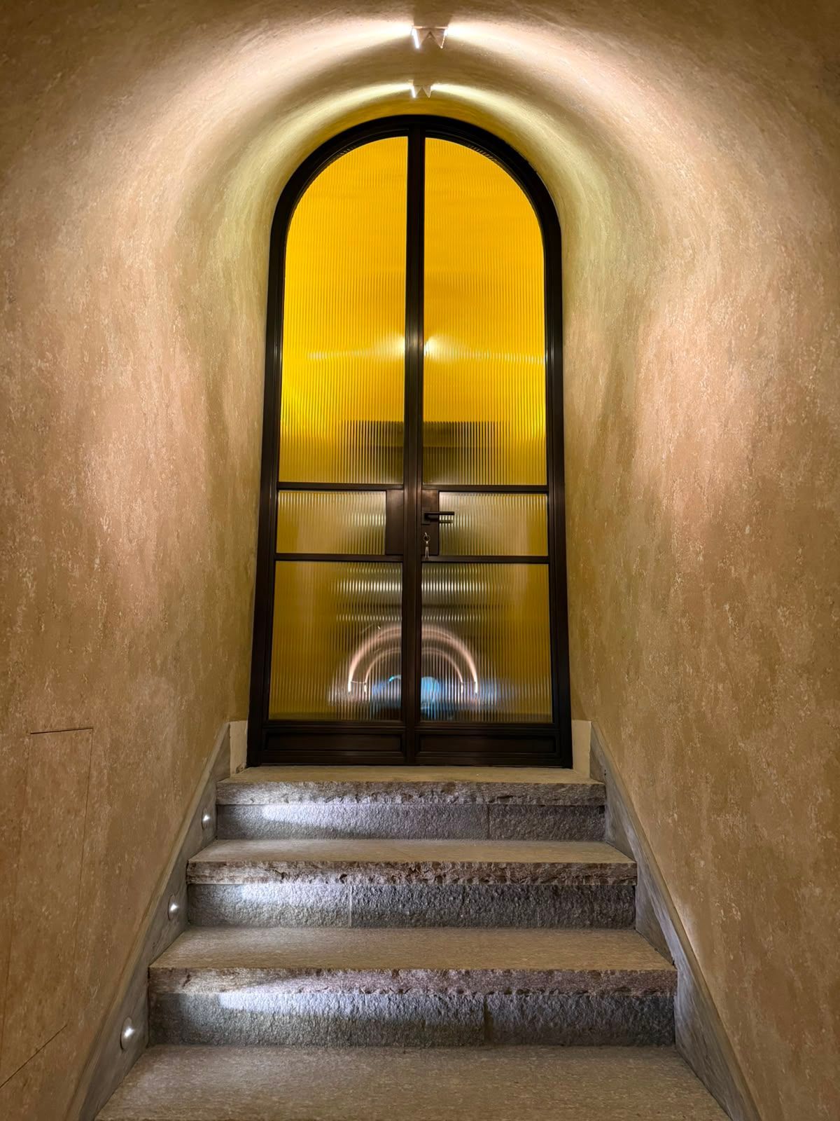 Una stretta scala conduce a una finestra ad arco illuminata di giallo, con cornice nera e pannelli di vetro.