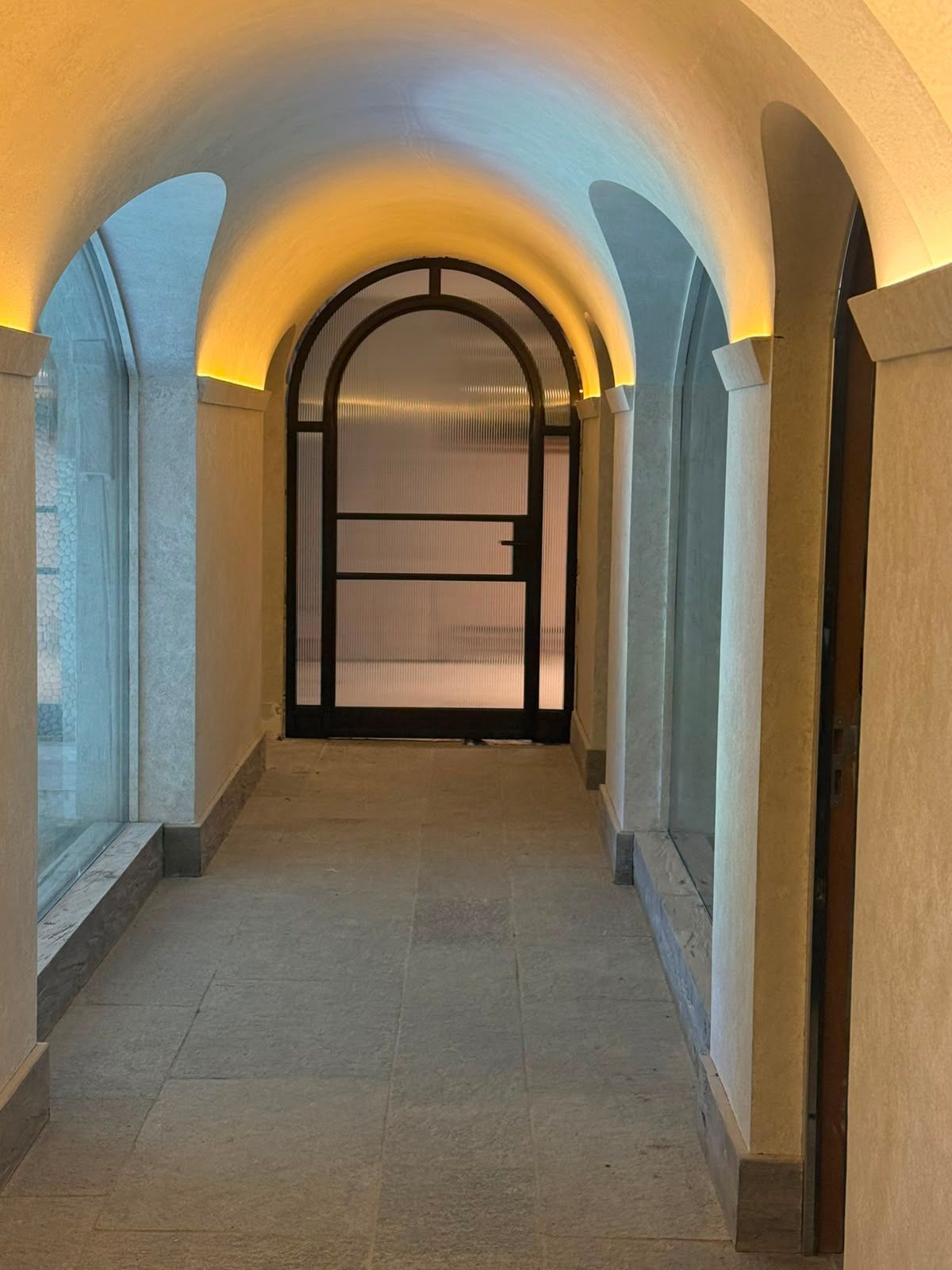 Corridoio ad arco con pavimento in pietra e illuminazione calda, che conduce a un cancello chiuso in fondo.