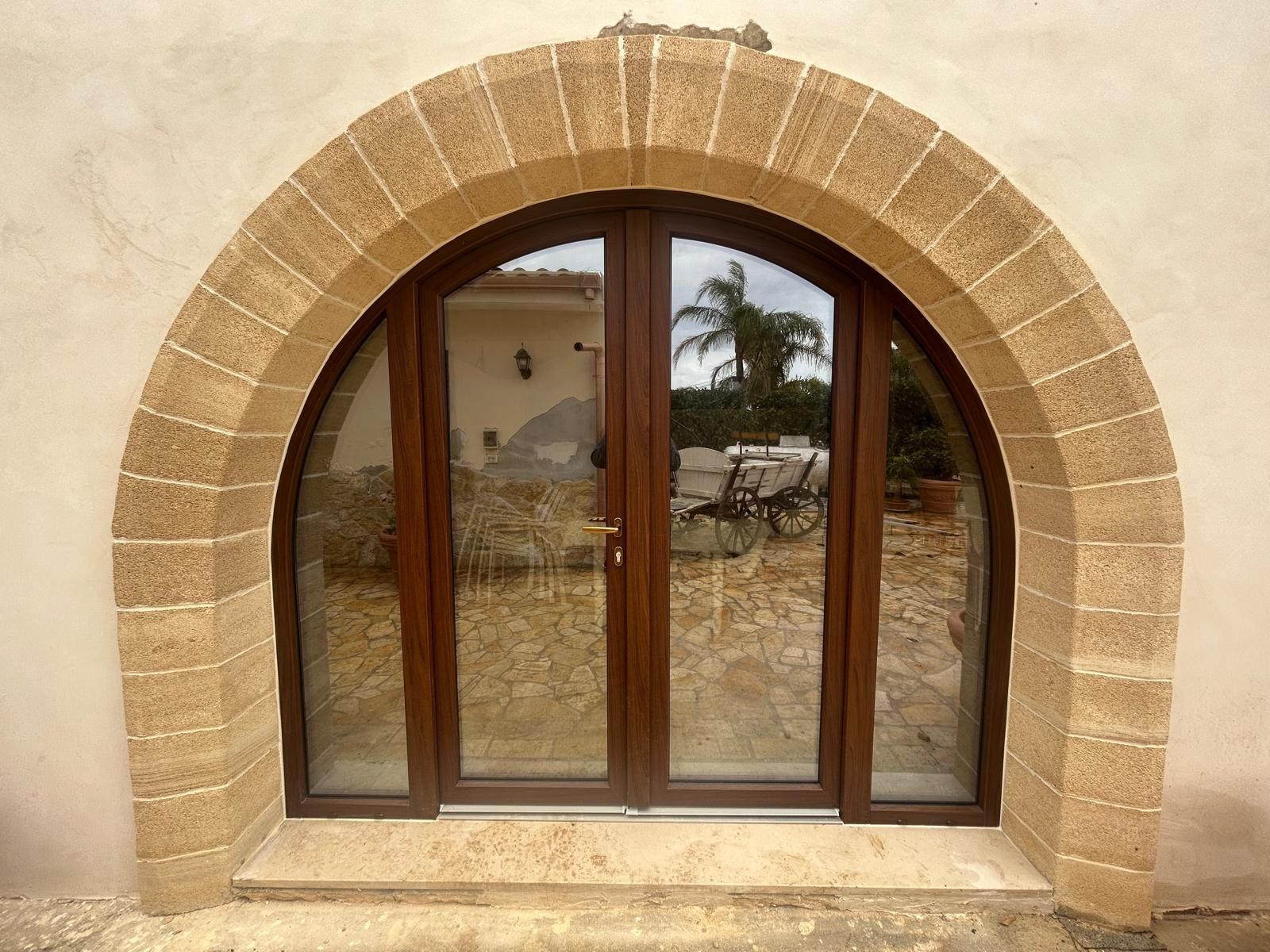 Arco in pietra che incornicia una porta a vetri con telaio in legno su una parete esterna intonacata.