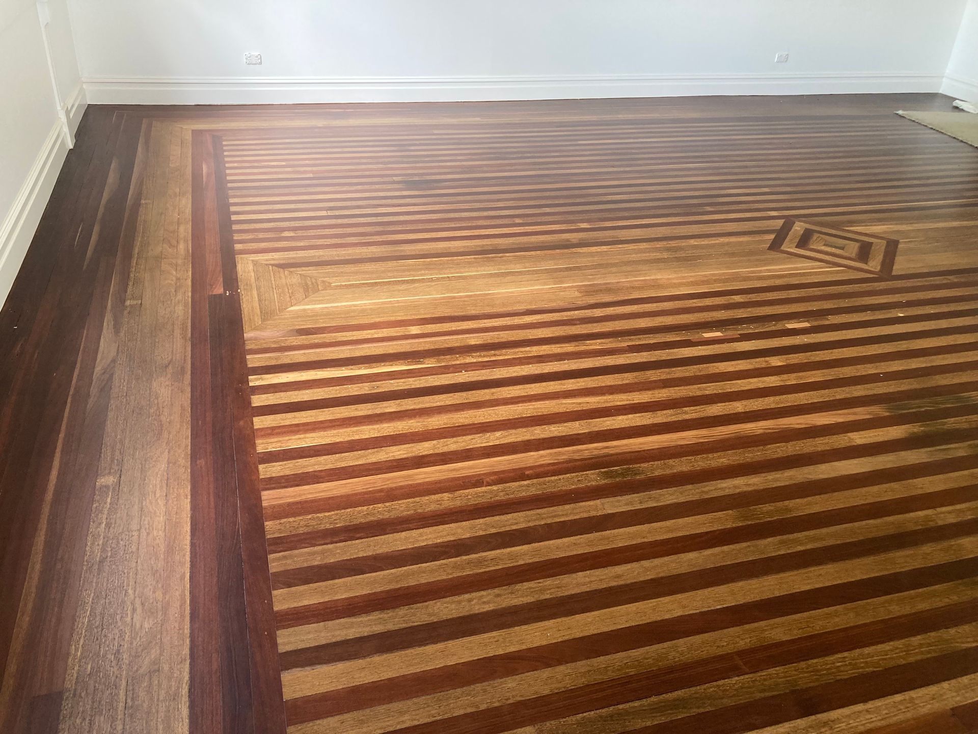 Wooden striped area rug on a hardwood floor in an empty room, near a white wall — Custom Floor Sanding In Somersby, NSW