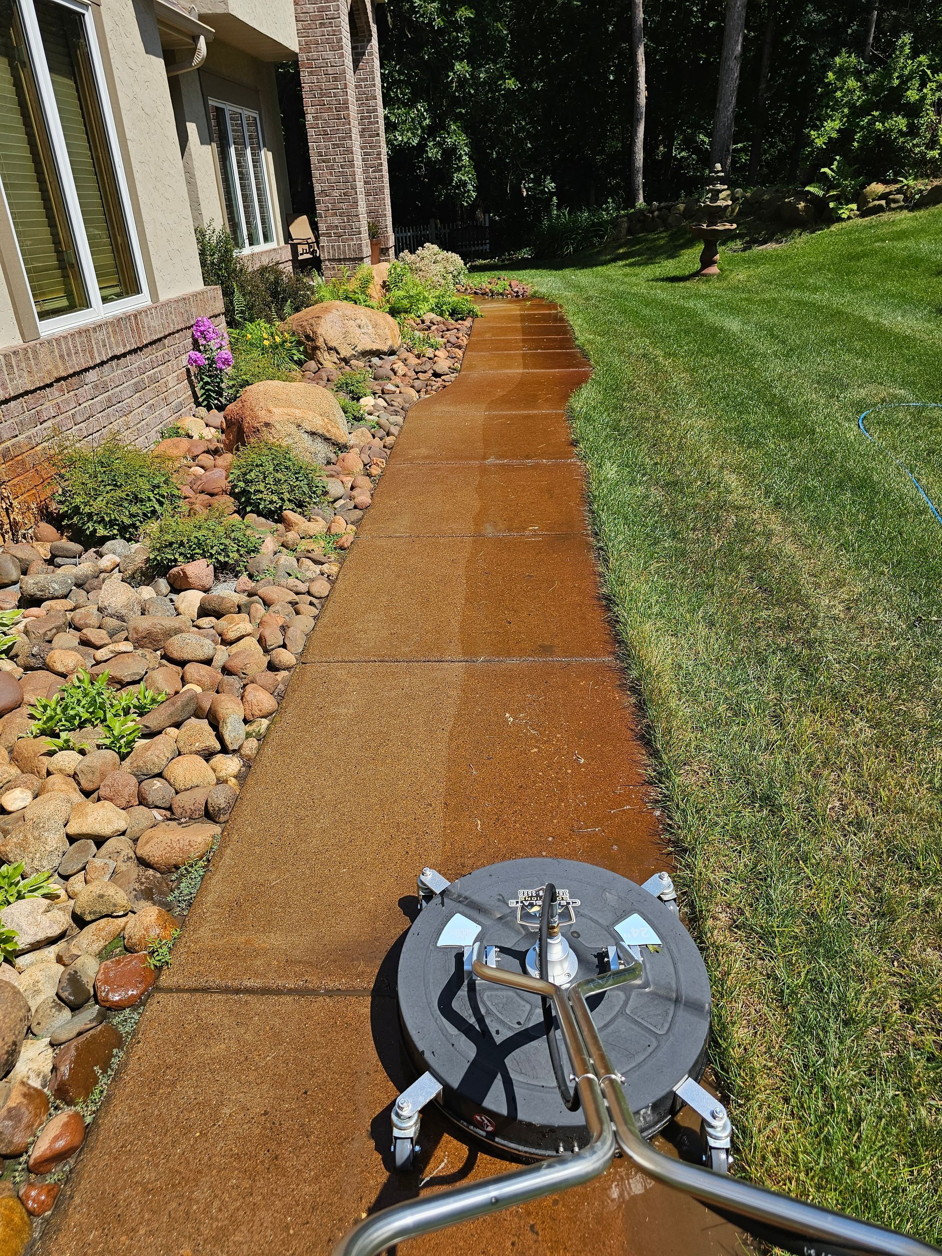 A surface cleaner moves along a dirty, rust-colored concrete sidewalk next to a house and a green lawn.