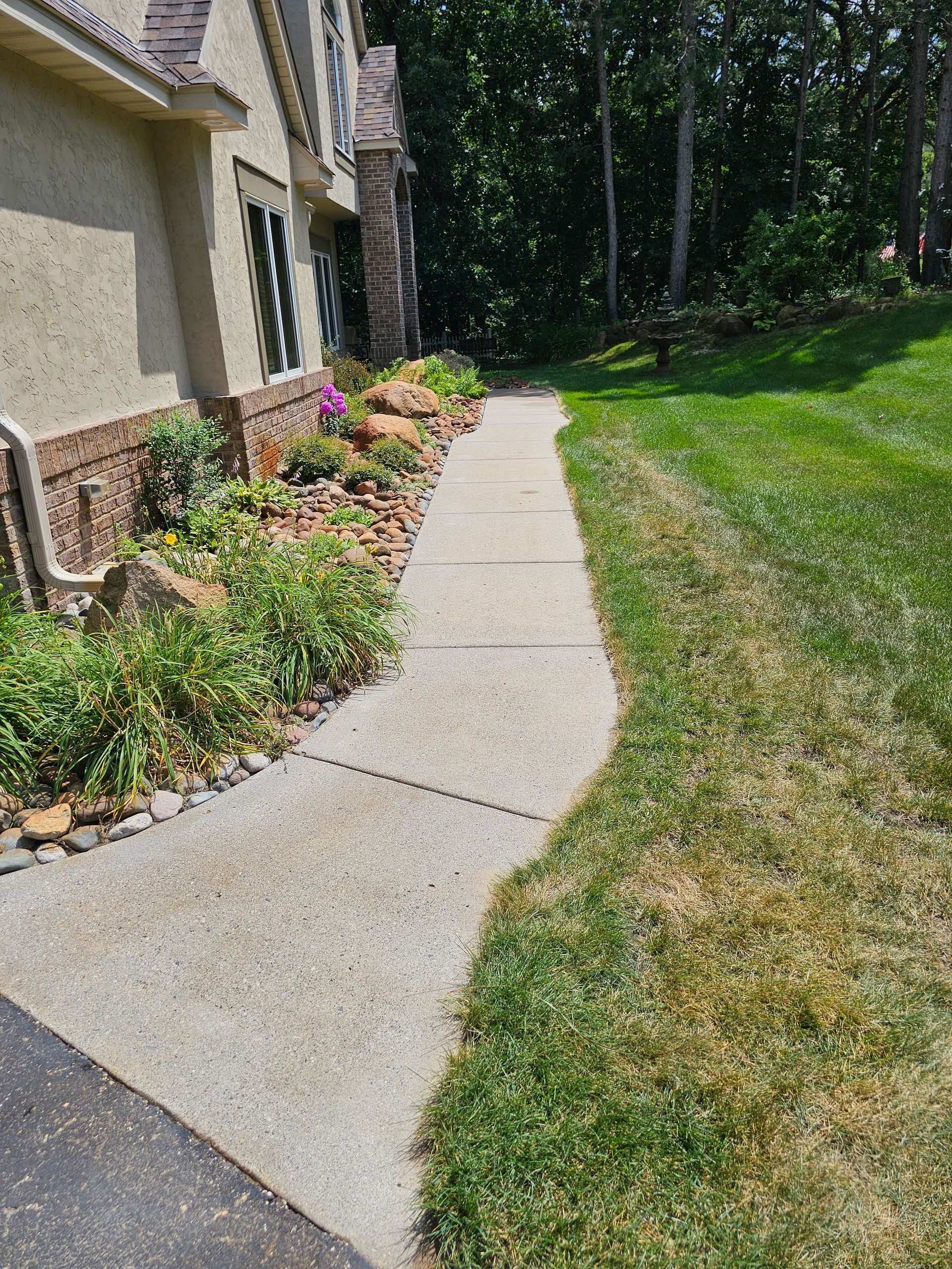 A concrete walkway leads along the side of a house past a landscaped garden bed and a grass lawn toward trees.
