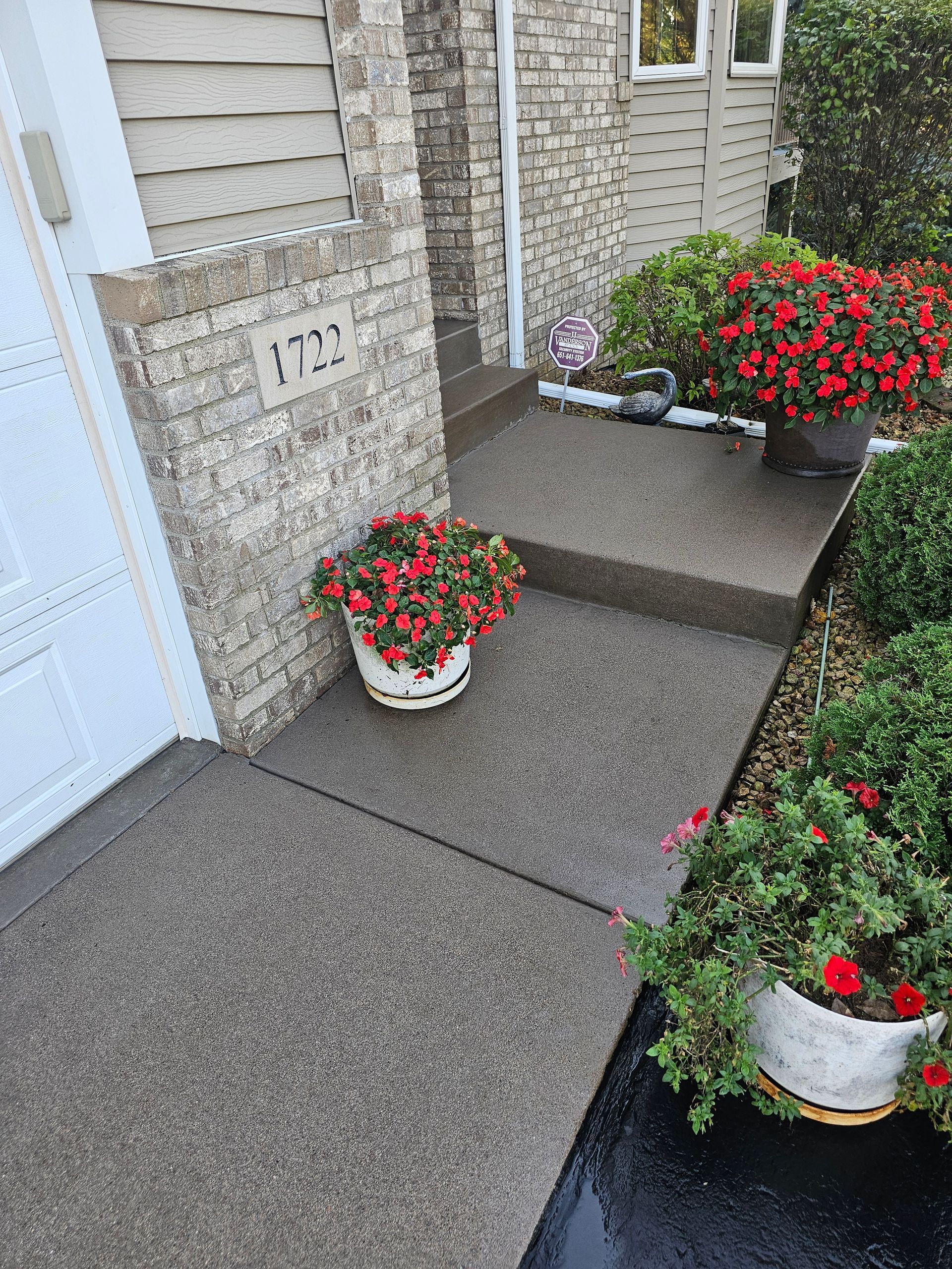 A porch entrance featuring concrete steps, brick exterior walls, and potted red flowers on a rainy day.