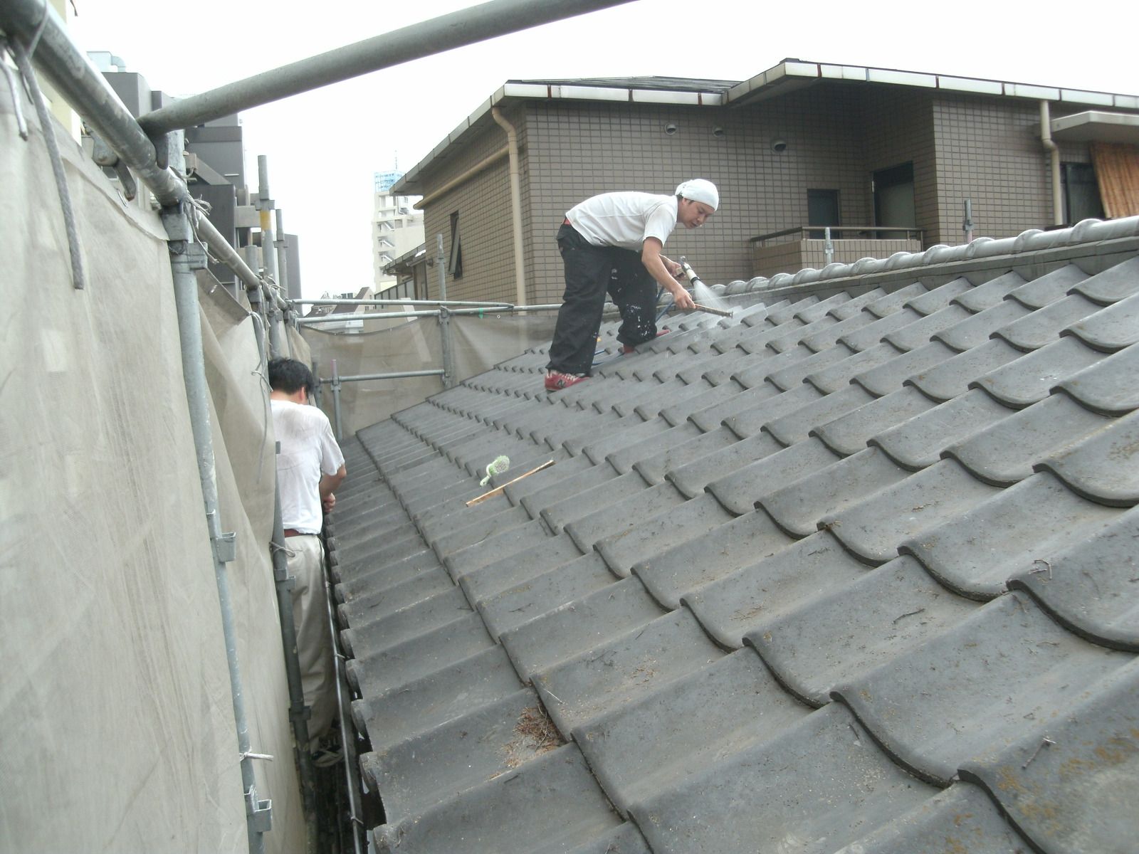 Two workers repairing a tiled roof; one sprays a substance while the other stands nearby.
