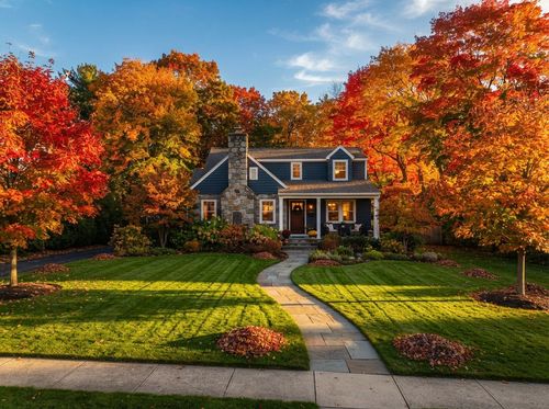 A suburban two-story house nestled among large, lush green trees with a manicured front lawn and a sunny, bright sky.