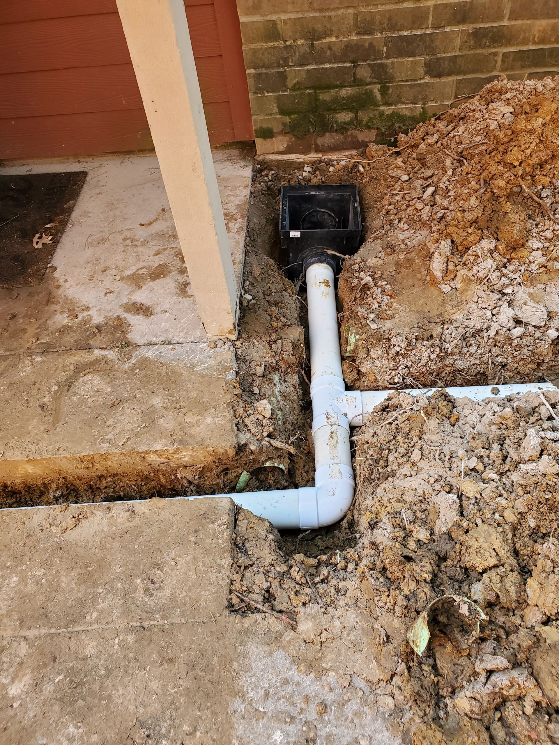 A gray PVC drainage pipe is installed in a trench next to a concrete patio and a brick wall.