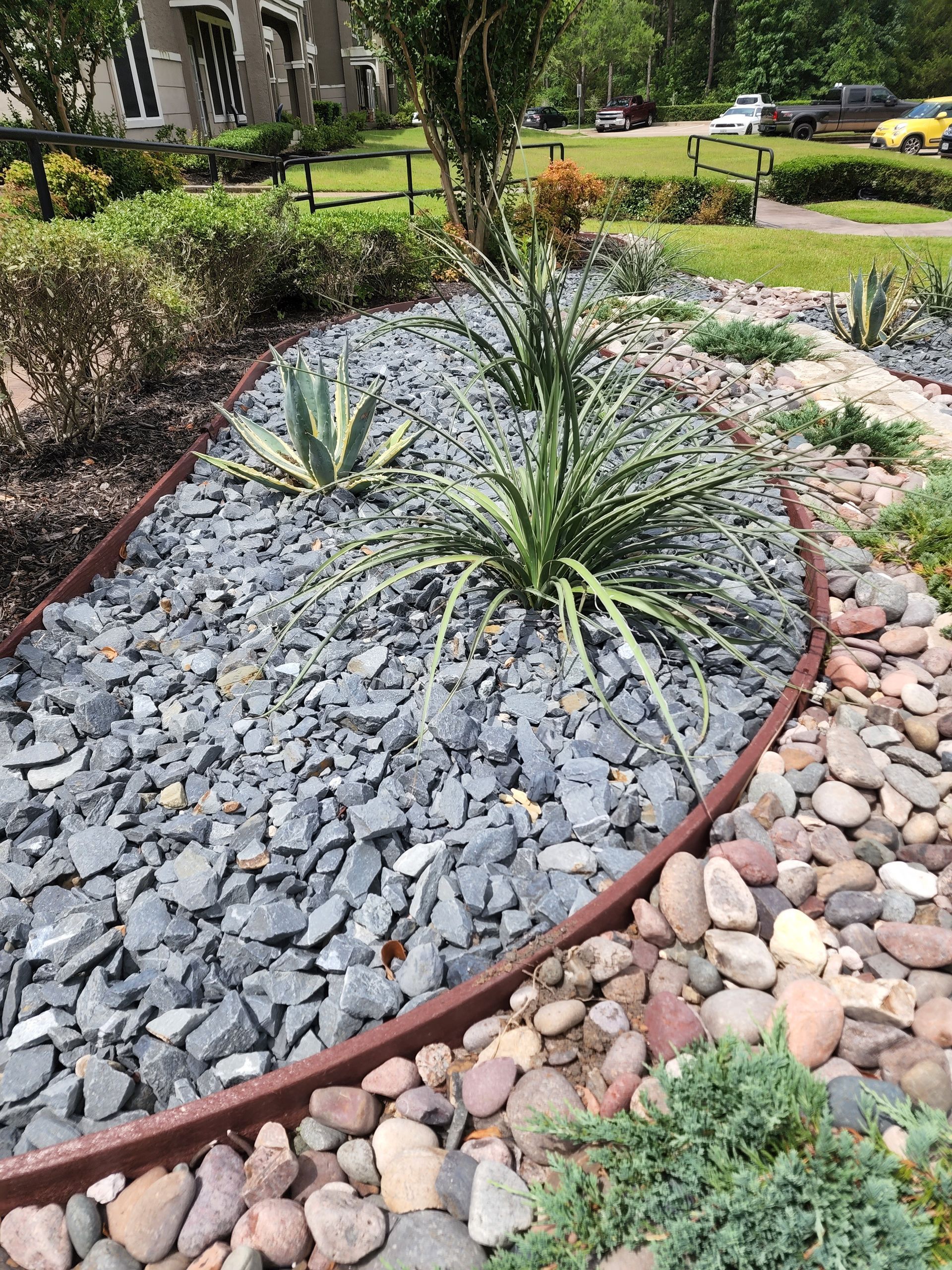 A garden bed features blue-gray gravel and agave plants bordered by a rusted metal edging next to a path of river rocks.