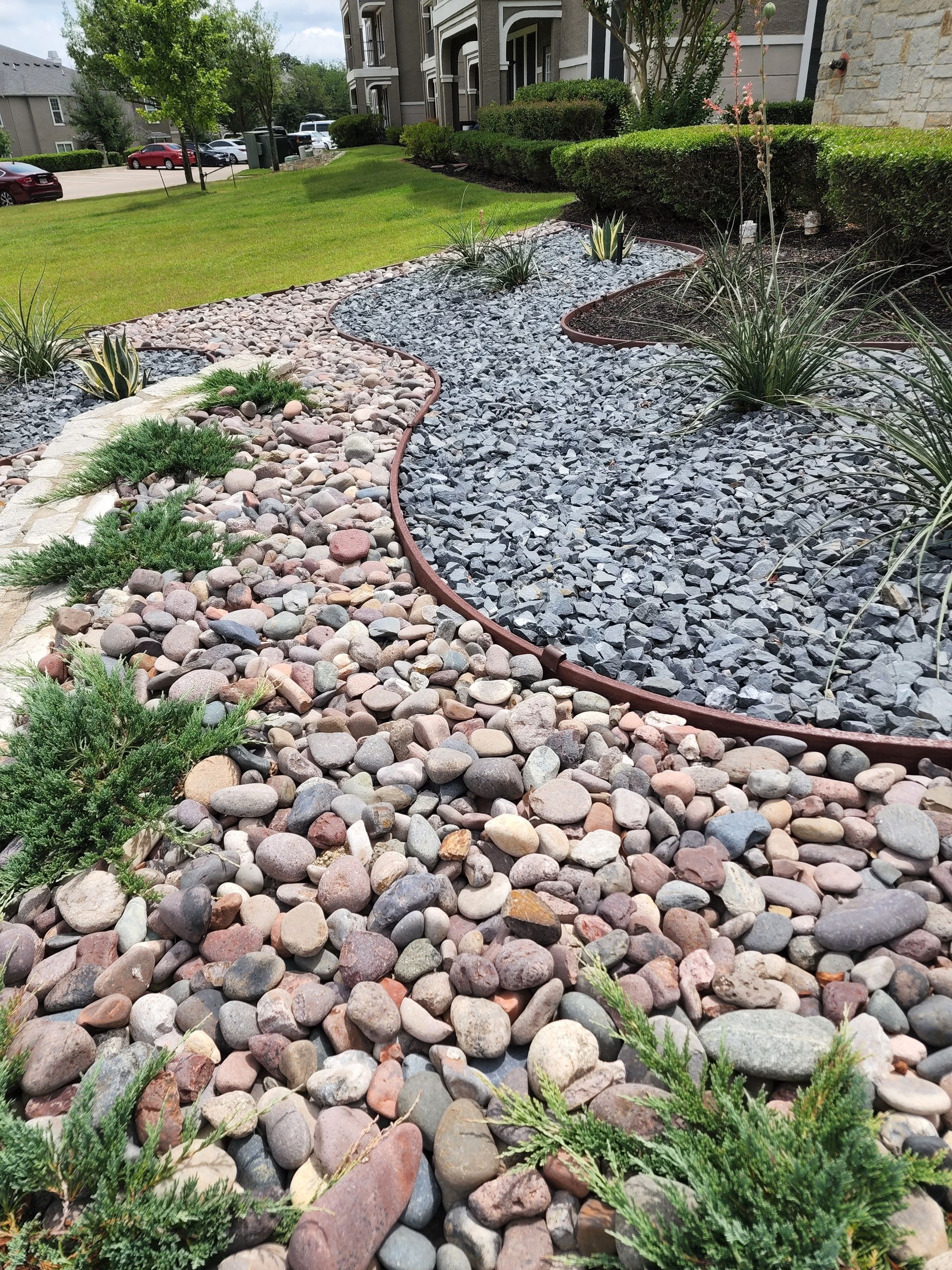 A landscaped garden bed featuring a curved border separating smooth, multicolored river rocks from dark gray gravel.