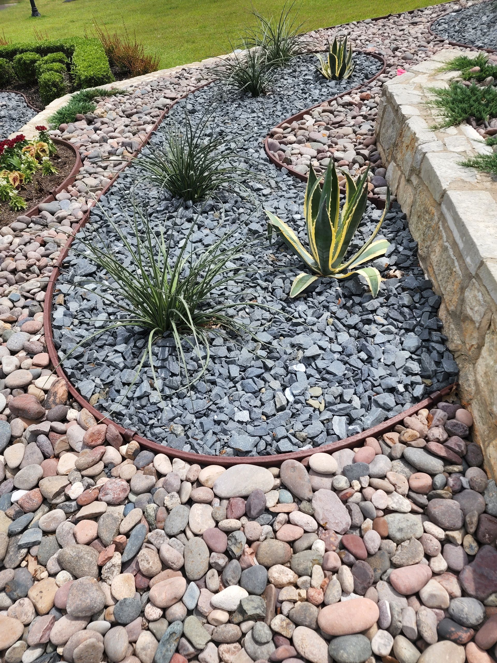 A curved garden bed of grey crushed stone featuring small succulents, bordered by a stone wall and larger river rocks.