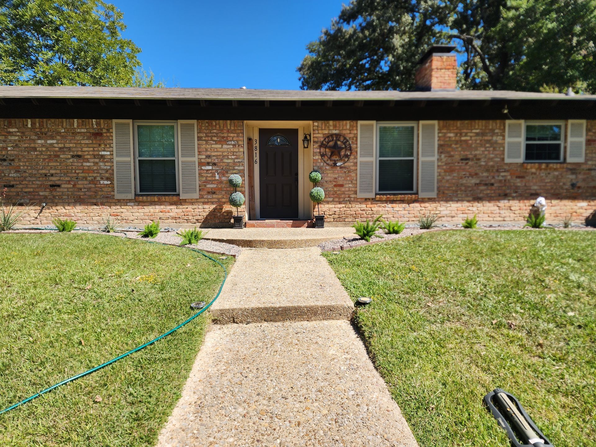 A single-story brick house with a concrete walkway leading to the front door, flanked by green lawn under a blue sky.