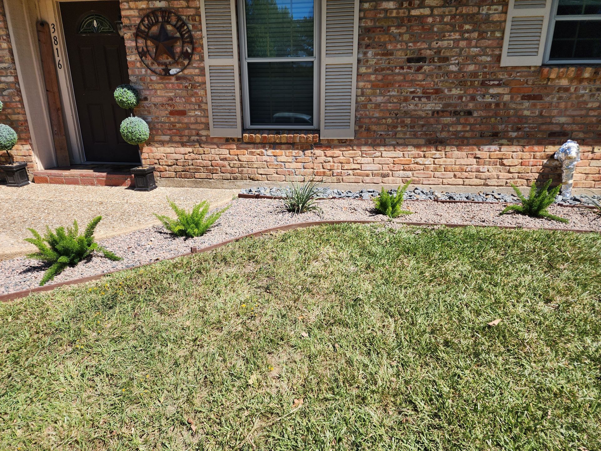 A brick house exterior with a mulched garden bed featuring small ferns and decorative rocks in front of a green lawn.