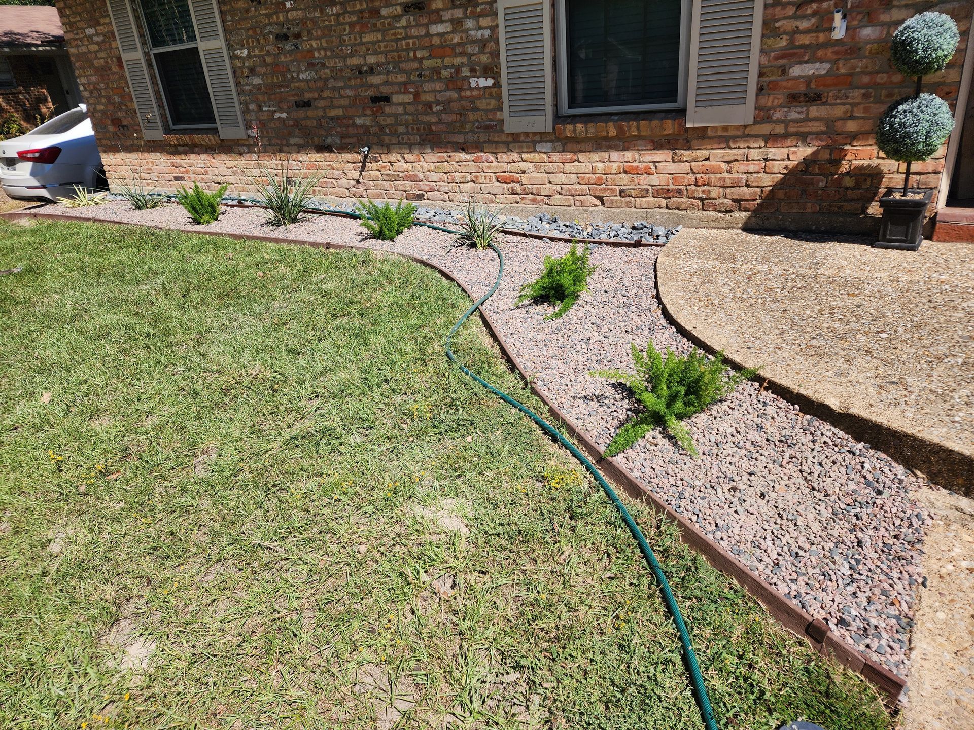 A brick house front with a curved garden bed of small purple gravel, green plants, and a green hose on the grass.