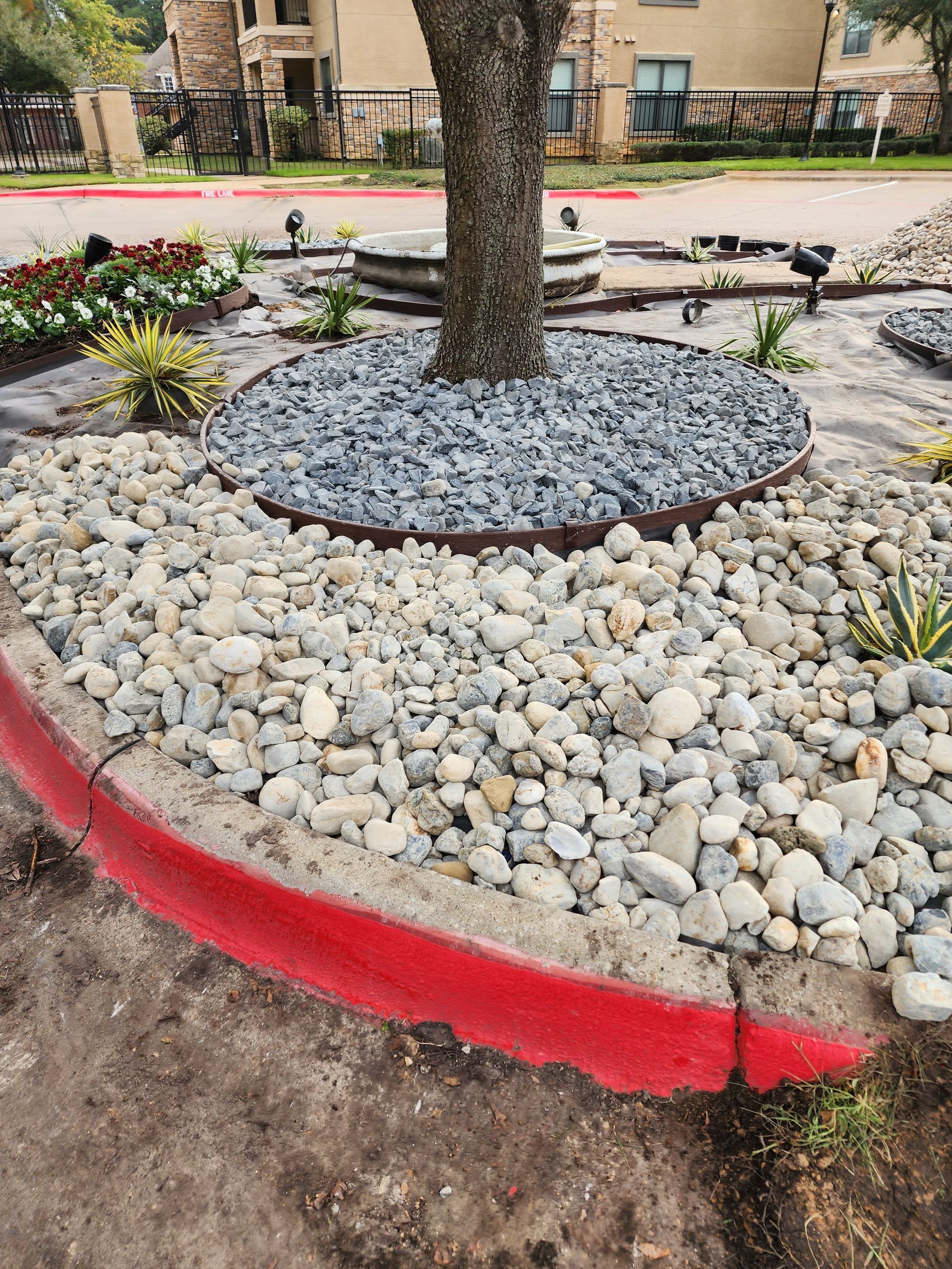 A tree island featuring a dark stone ring, river rocks, and a red concrete curb in a parking lot.