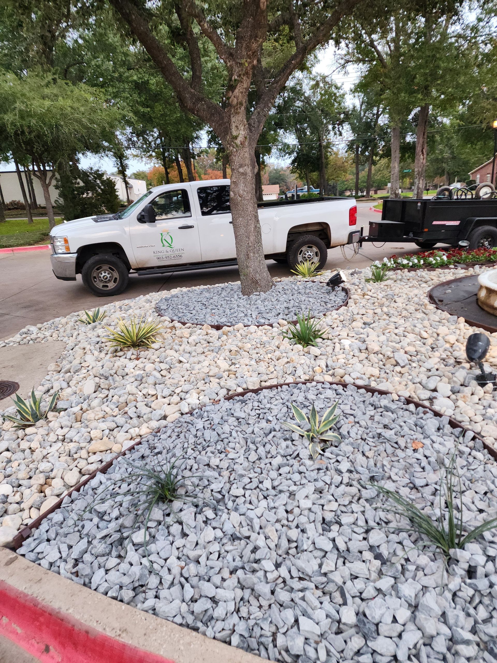 Landscaping scene showing a white pickup truck, a utility trailer, and new beds of white and gray stone mulch.