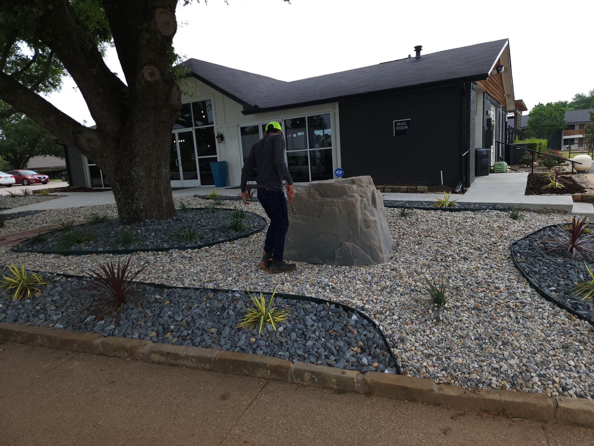 A person in a hard hat stands near a large boulder in a landscaped yard with decorative gravel and a building behind.