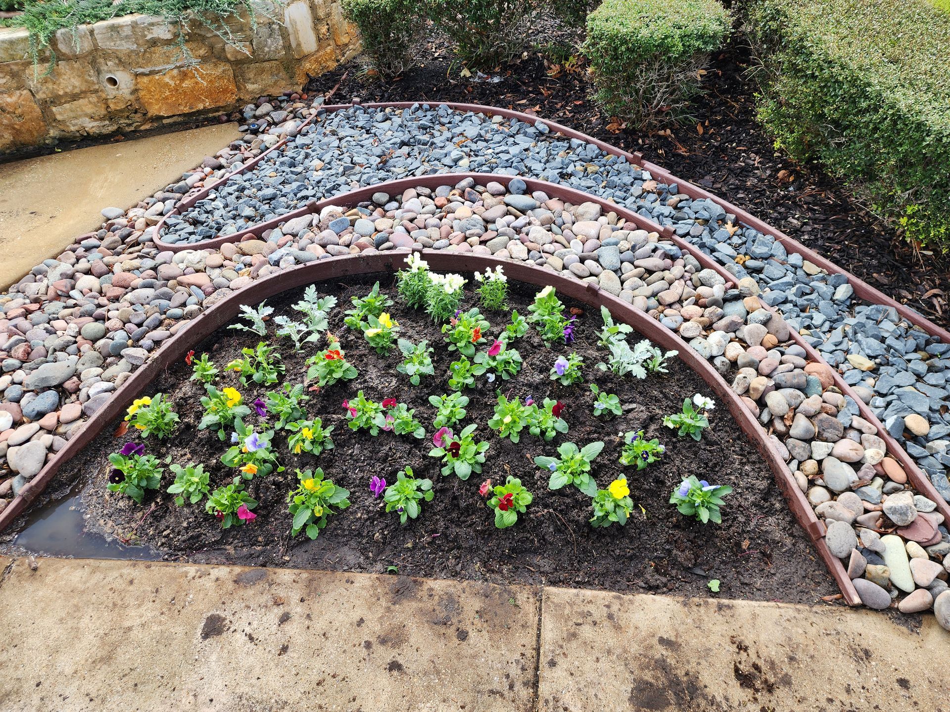 A semi-circular garden bed with colorful flowers, bordered by curved landscaping metal and rows of smooth river stones.