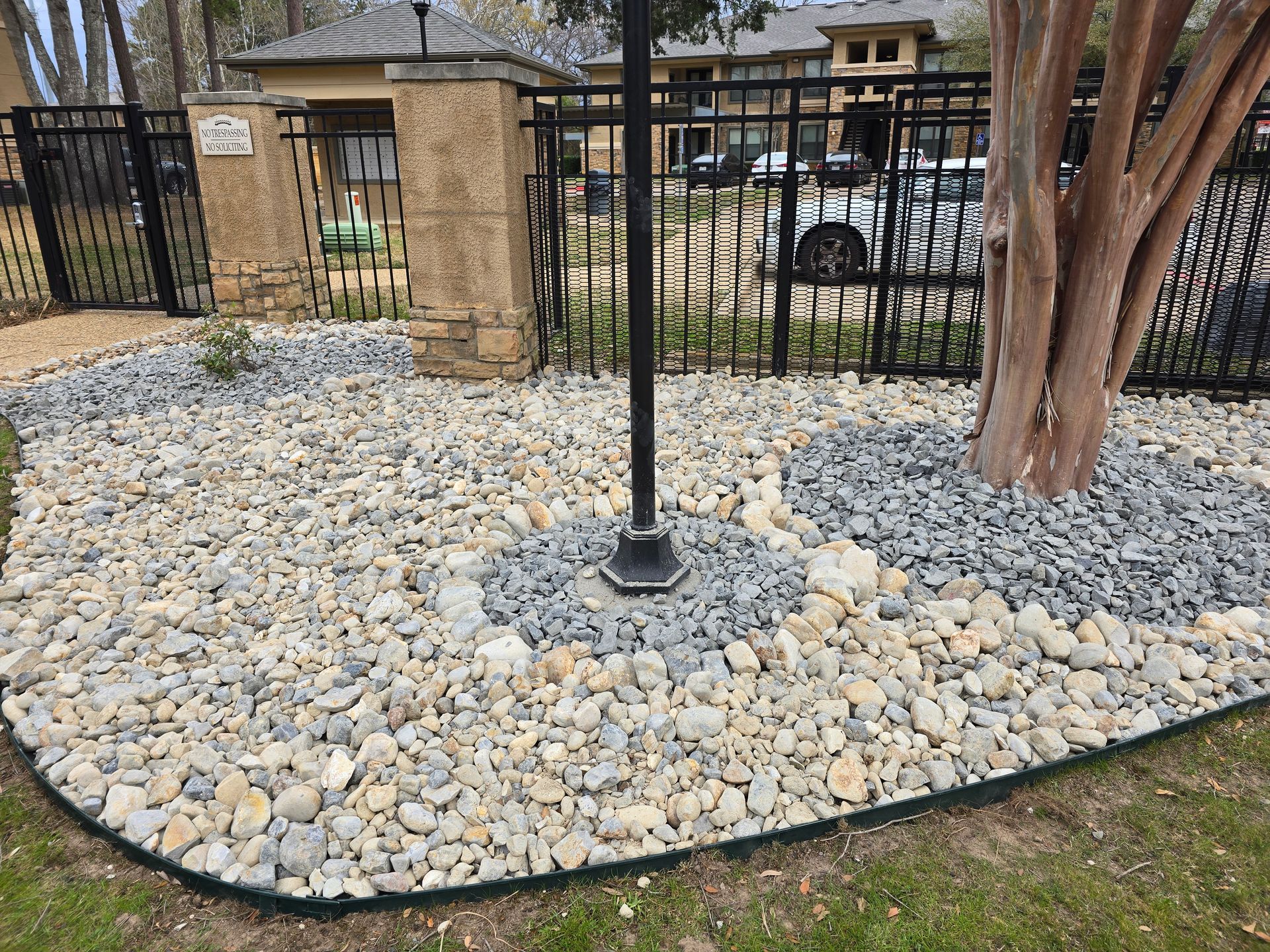 A landscaping bed features a mix of light tan and grey rocks, encircling a tree and a black metal pole by a fenced gate.