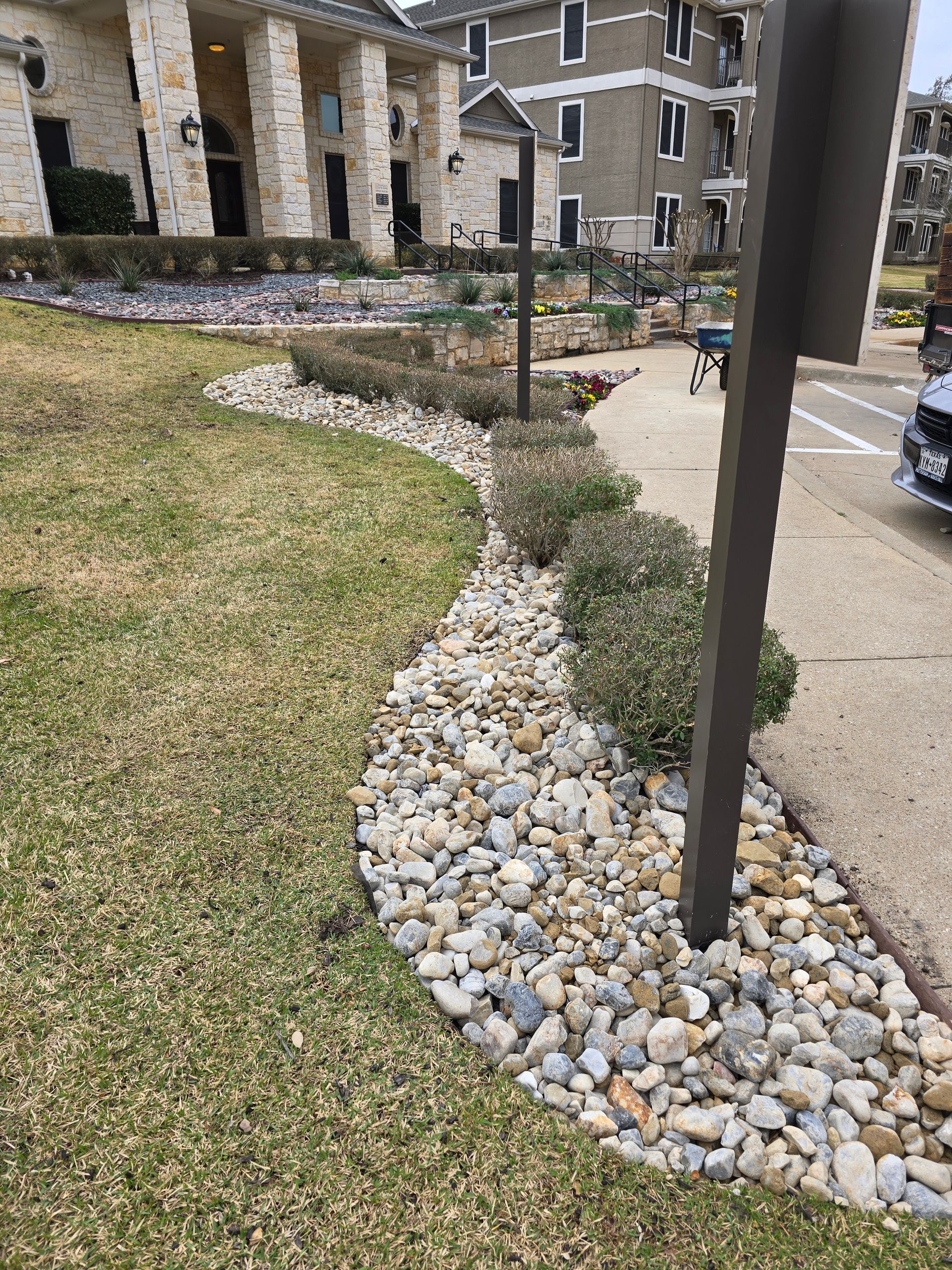 Landscaped rock garden bed with shrubs bordering a paved driveway near a stone building.