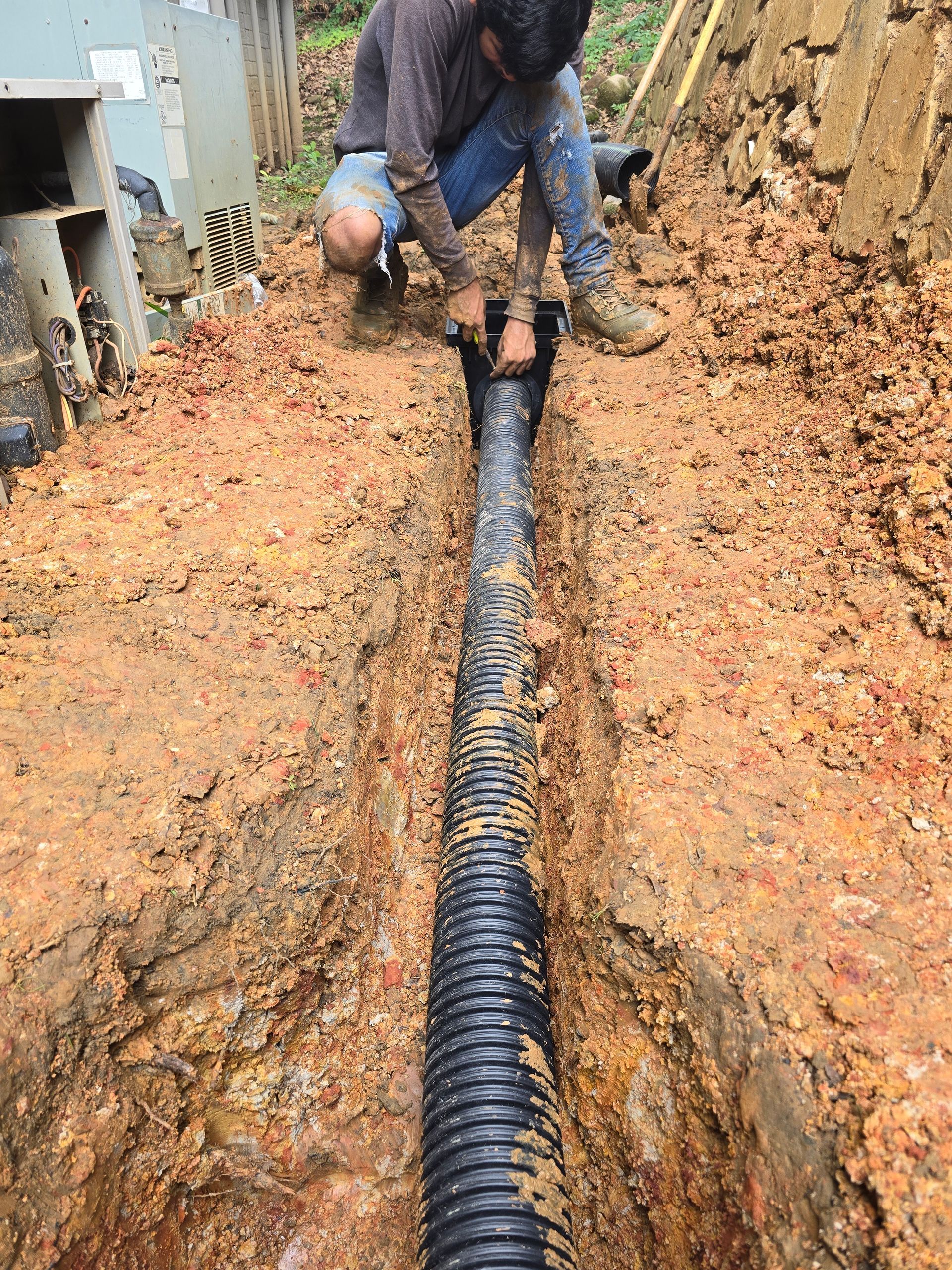 A person kneels in a muddy trench, connecting a section of black corrugated drainage pipe.