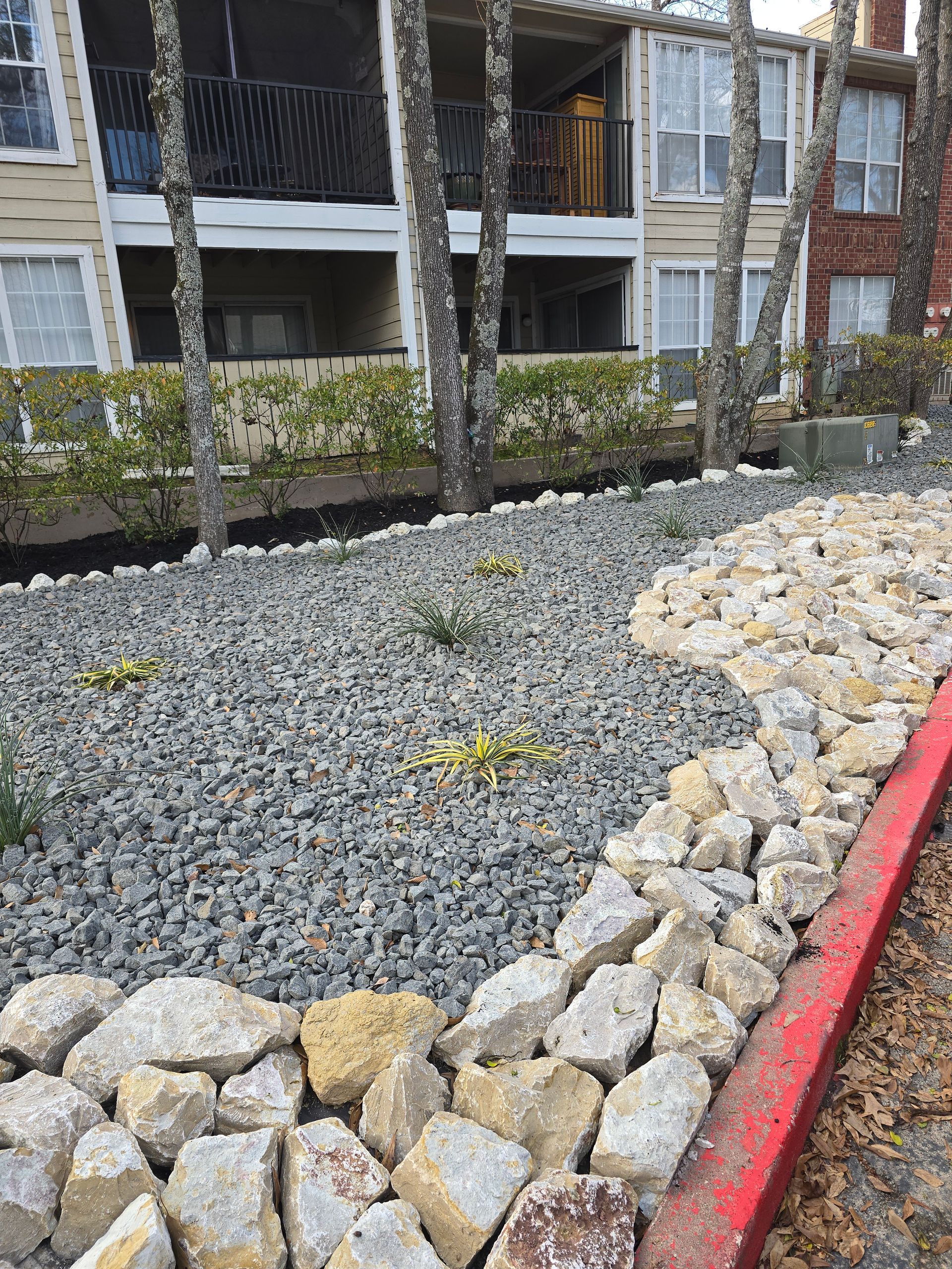 A landscaped area with gray gravel, a border of light-colored rocks, and a red curb in front of an apartment building.