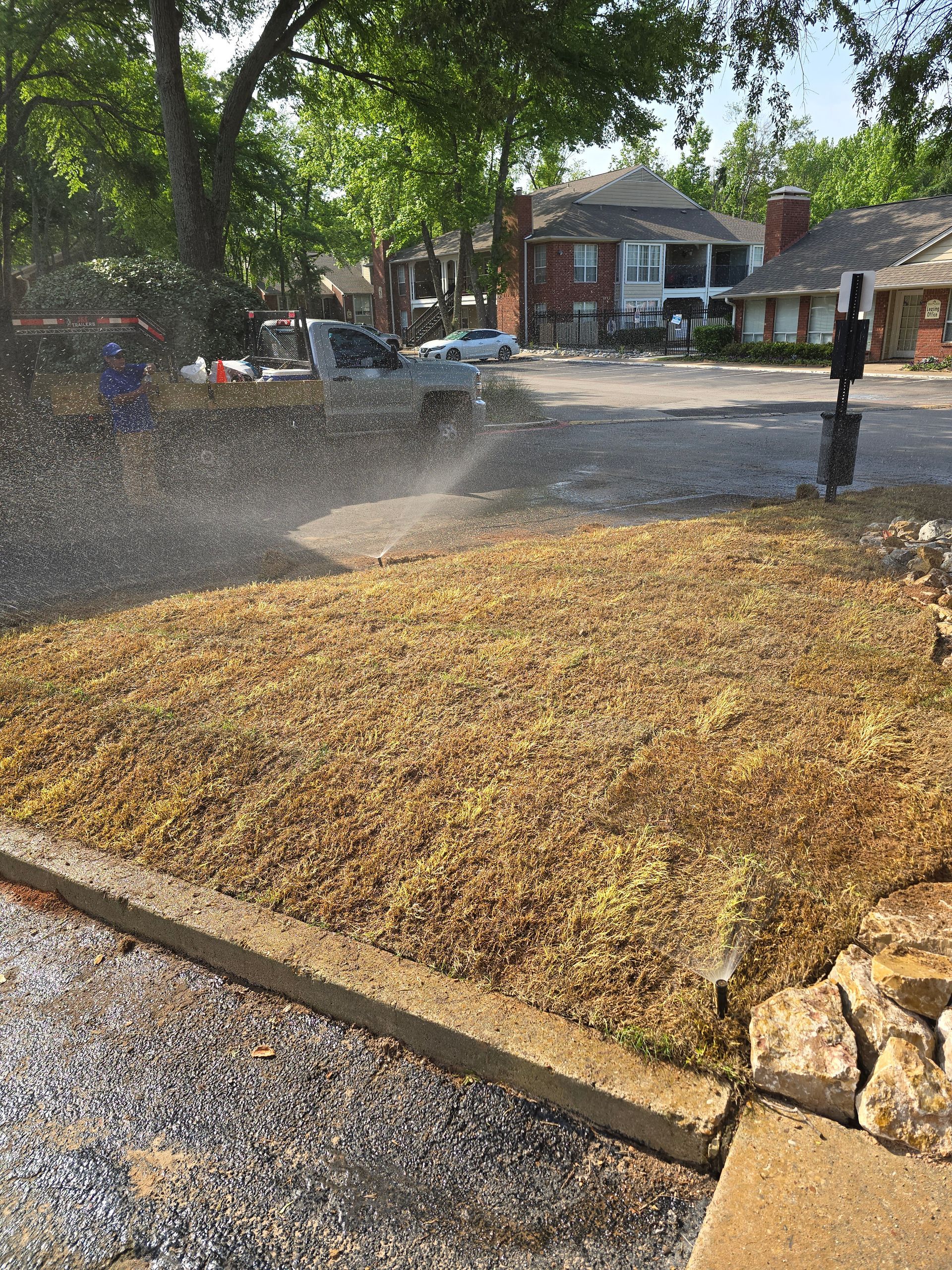 A sprinkler waters a patch of freshly mulched dirt and gravel in front of a residential apartment building.