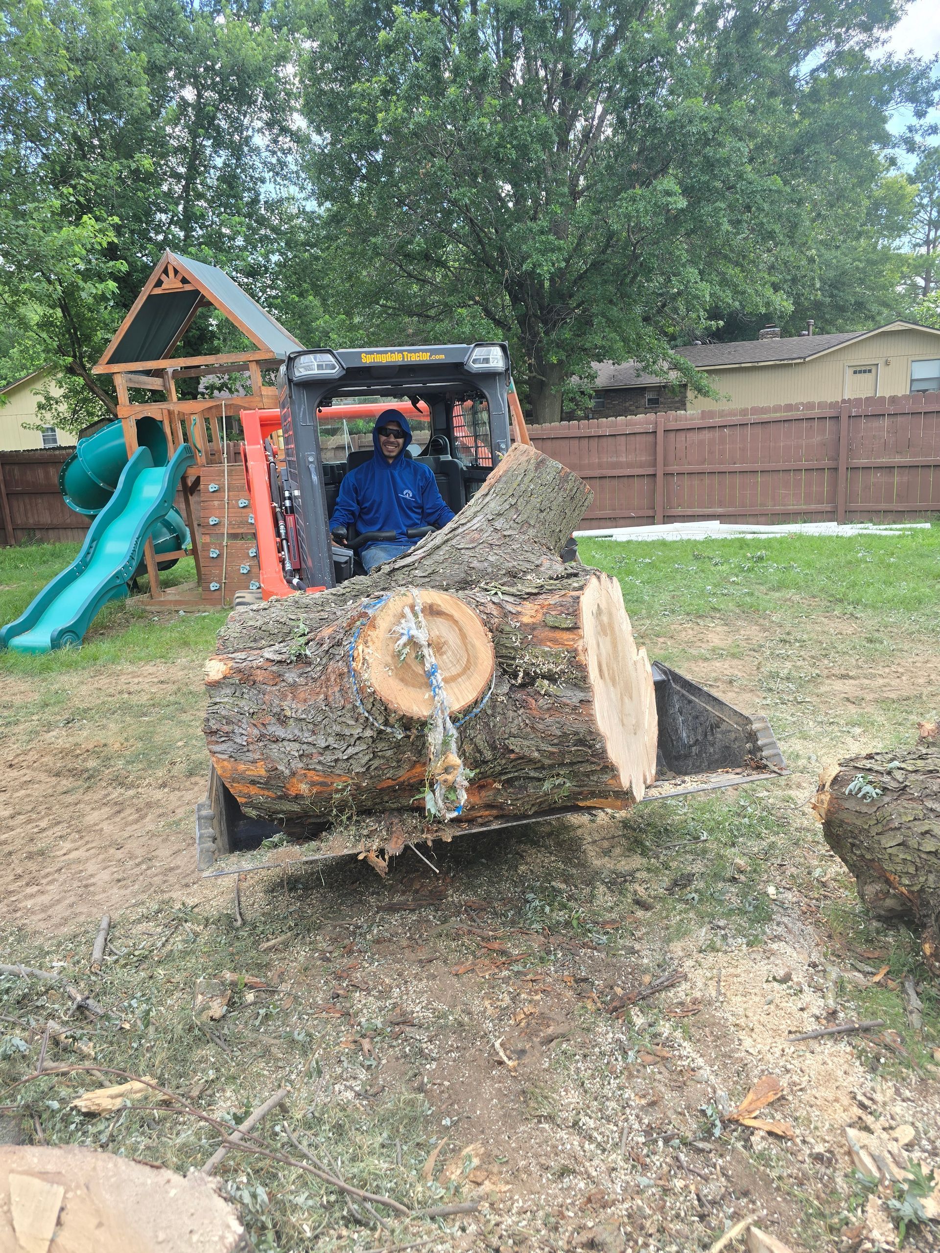 A worker operating a skid steer to lift a large tree log in a residential backyard with a play structure nearby.