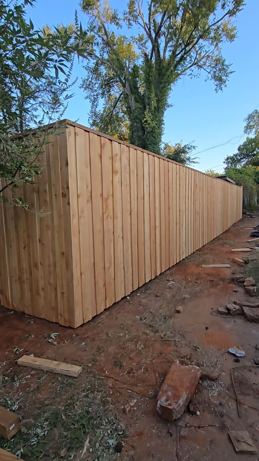 A new, tall, light-brown wood privacy fence stretches across a yard with dirt ground under a clear blue sky.