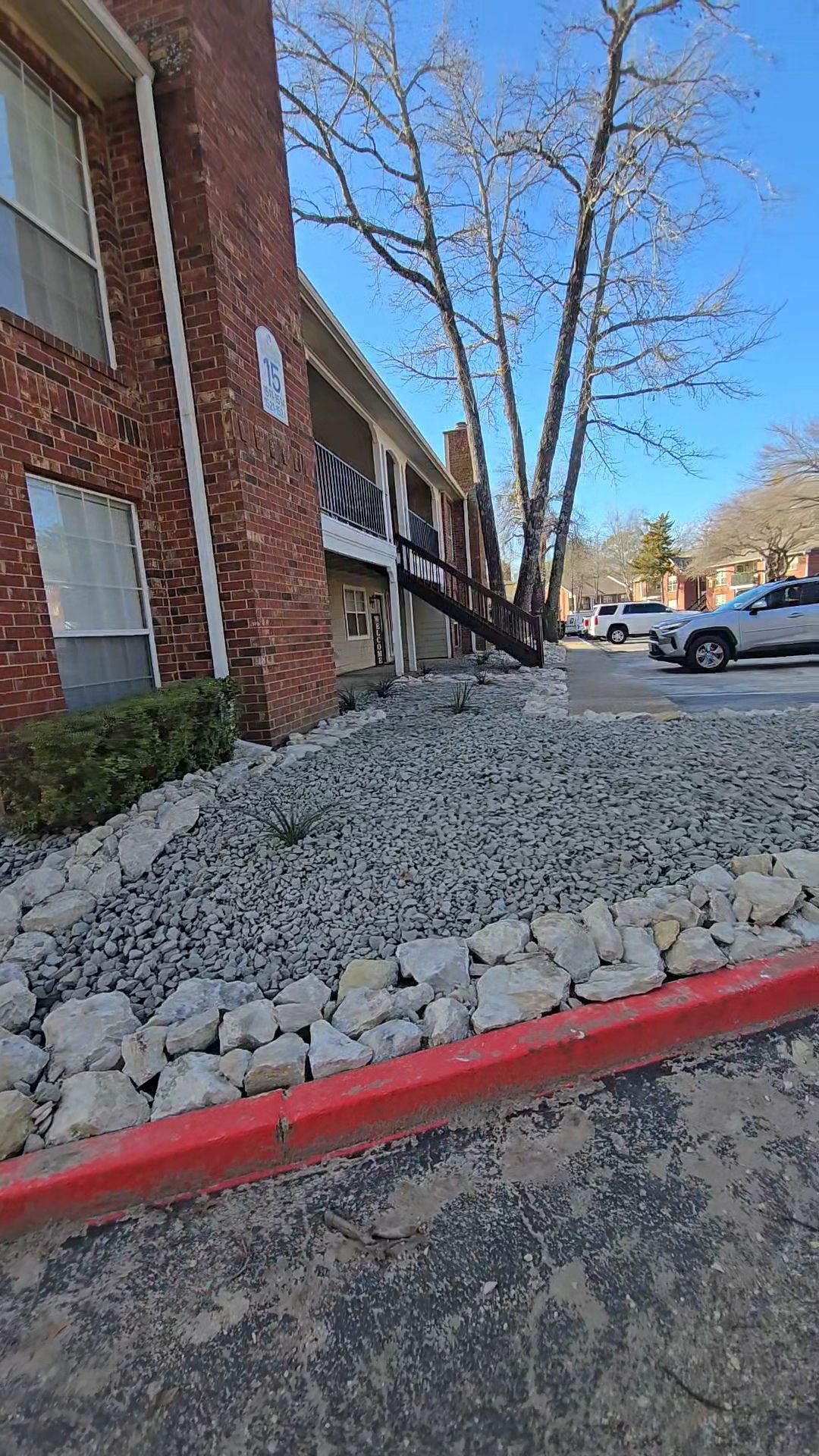 A brick apartment building with exterior staircases, a rocky landscape bed, and a red curb in a parking lot.