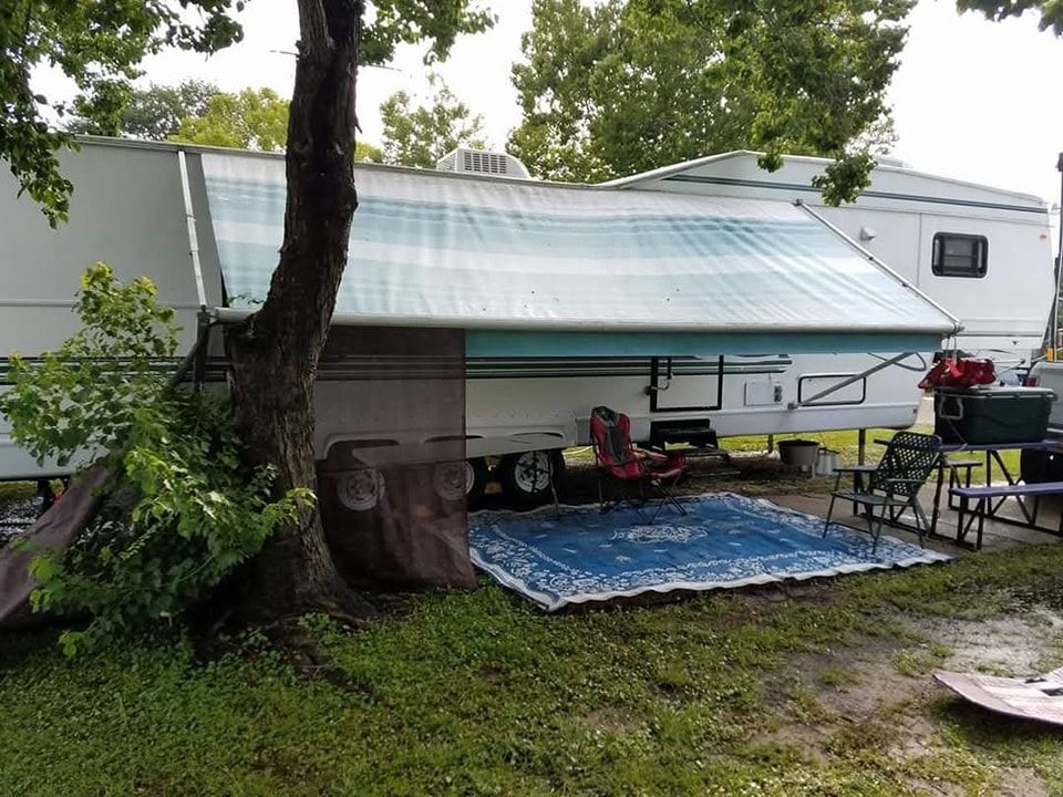 White RV with teal awning beside a tree, covering a small campsite with chairs and a blue rug