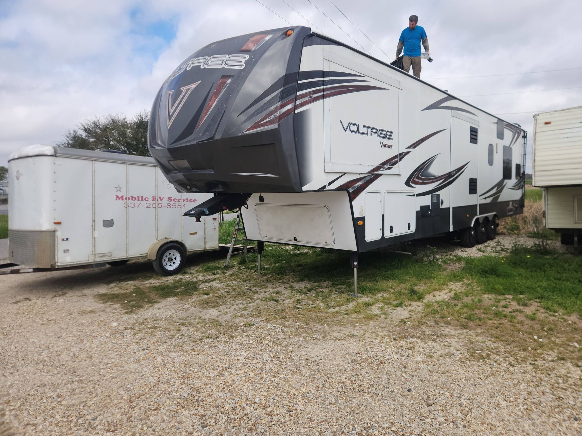 Large fifth-wheel RV parked on gravel, with a person standing on the roof and trailers nearby.