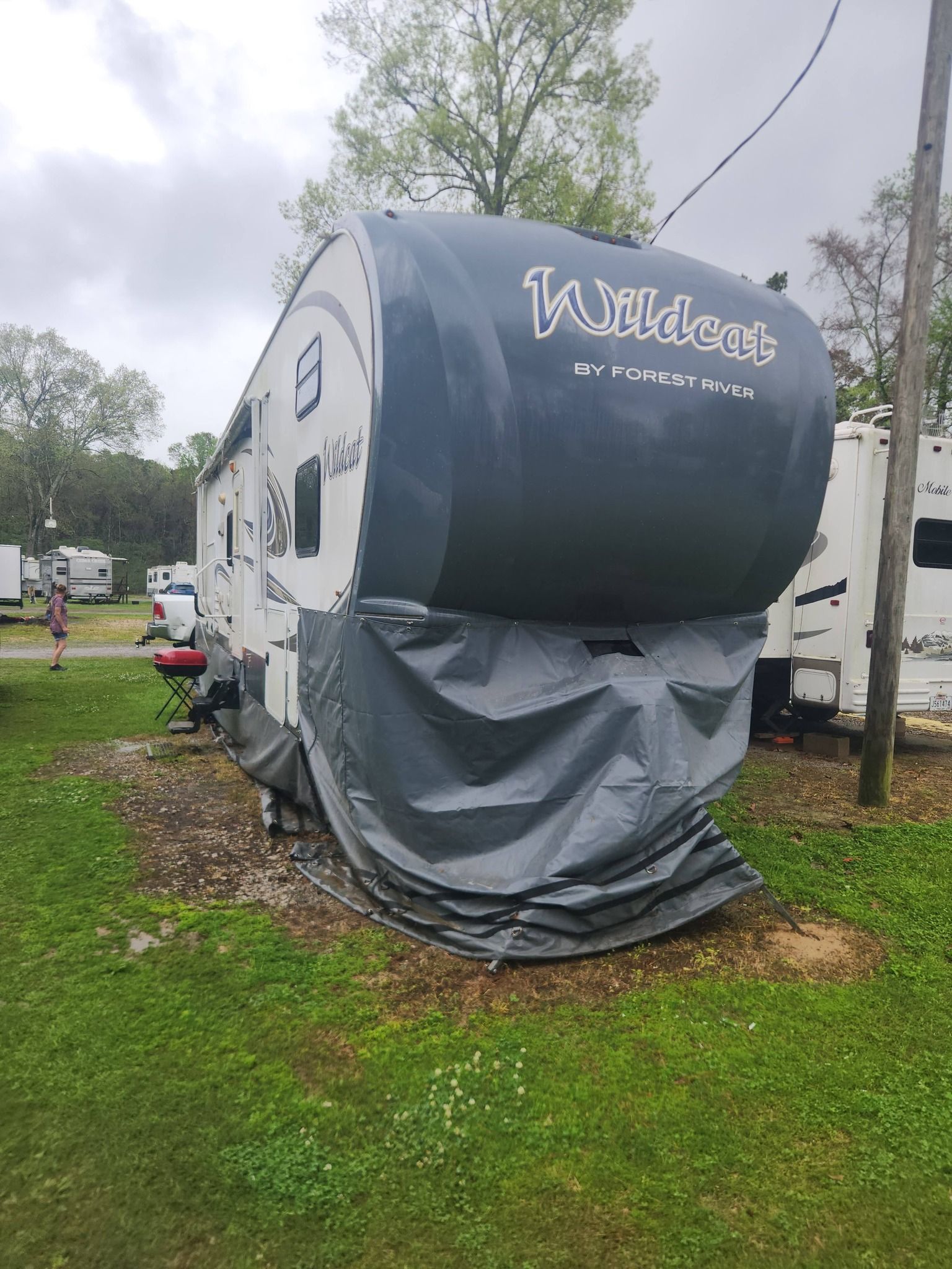 Black and gray travel trailer with front cover and gray skirt parked on grass at a campground