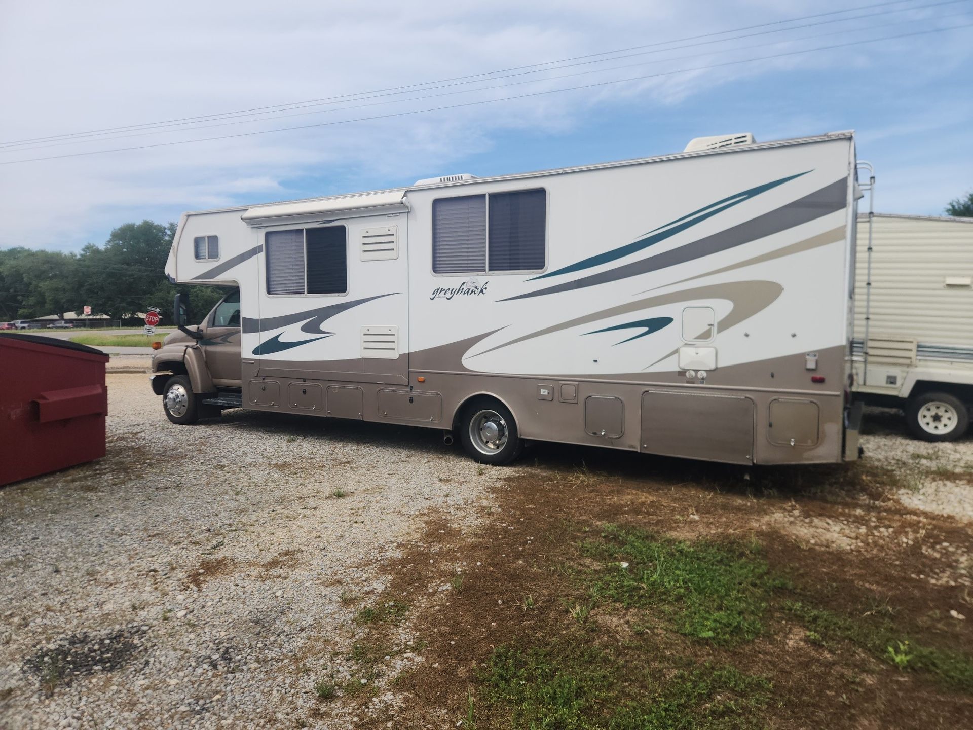 White and beige RV camper parked on gravel beside a red dumpster under a cloudy sky