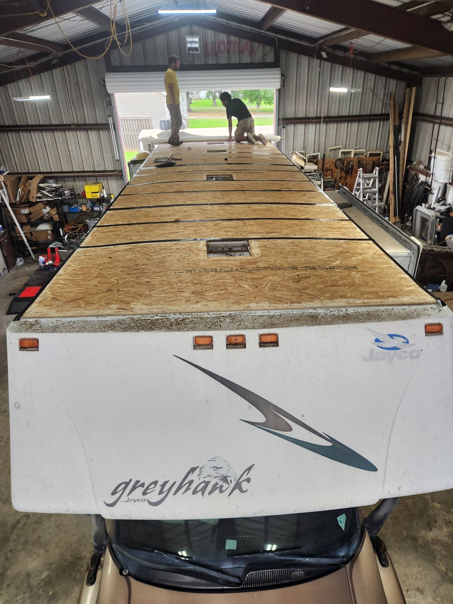 Inside a shed, two workers stand on a large rooftop camper trailer with a plywood-covered roof.