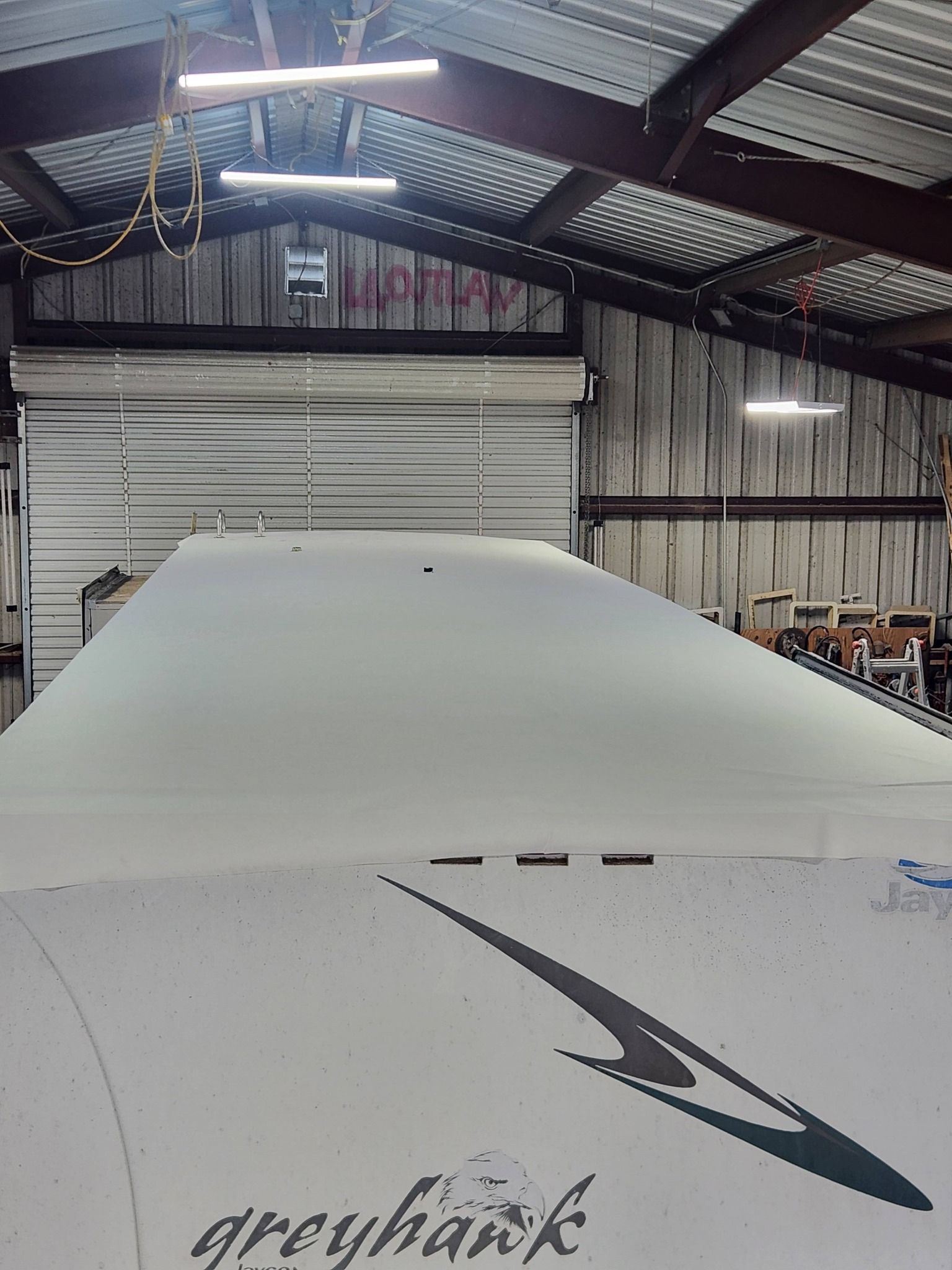 Inside a metal garage, the nose of a white Greyhawk RV under bright overhead lights