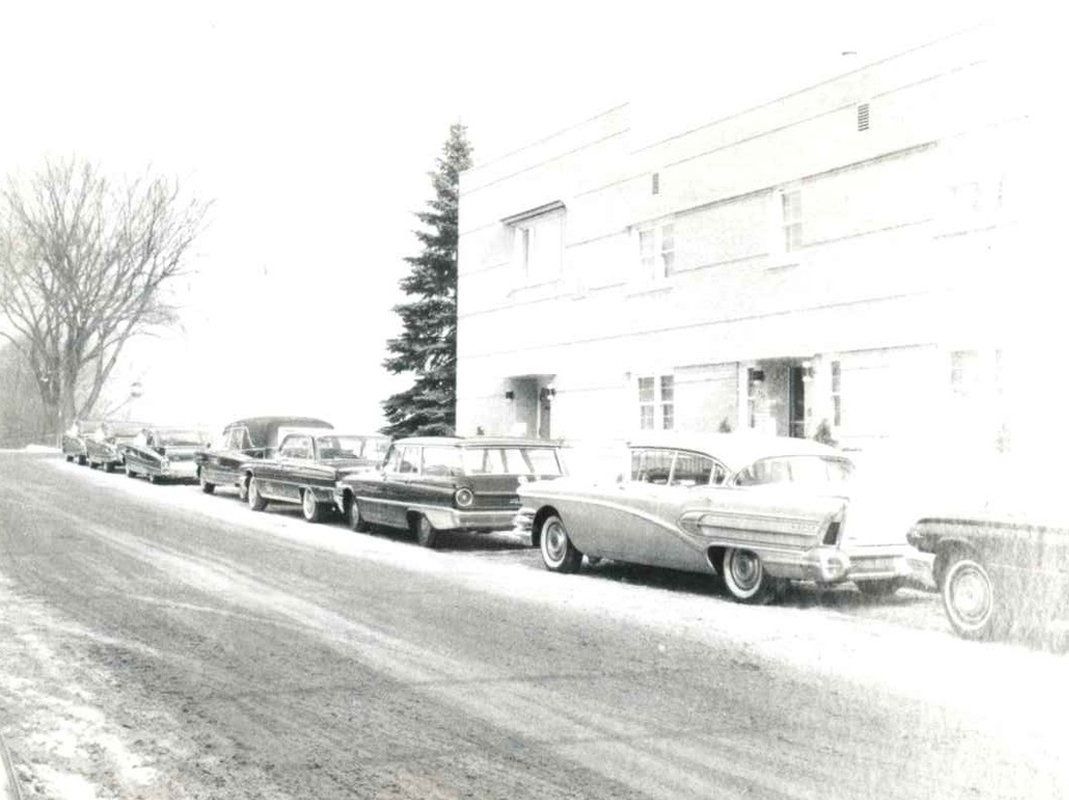A black and white photo of a row of cars parked in front of a building.