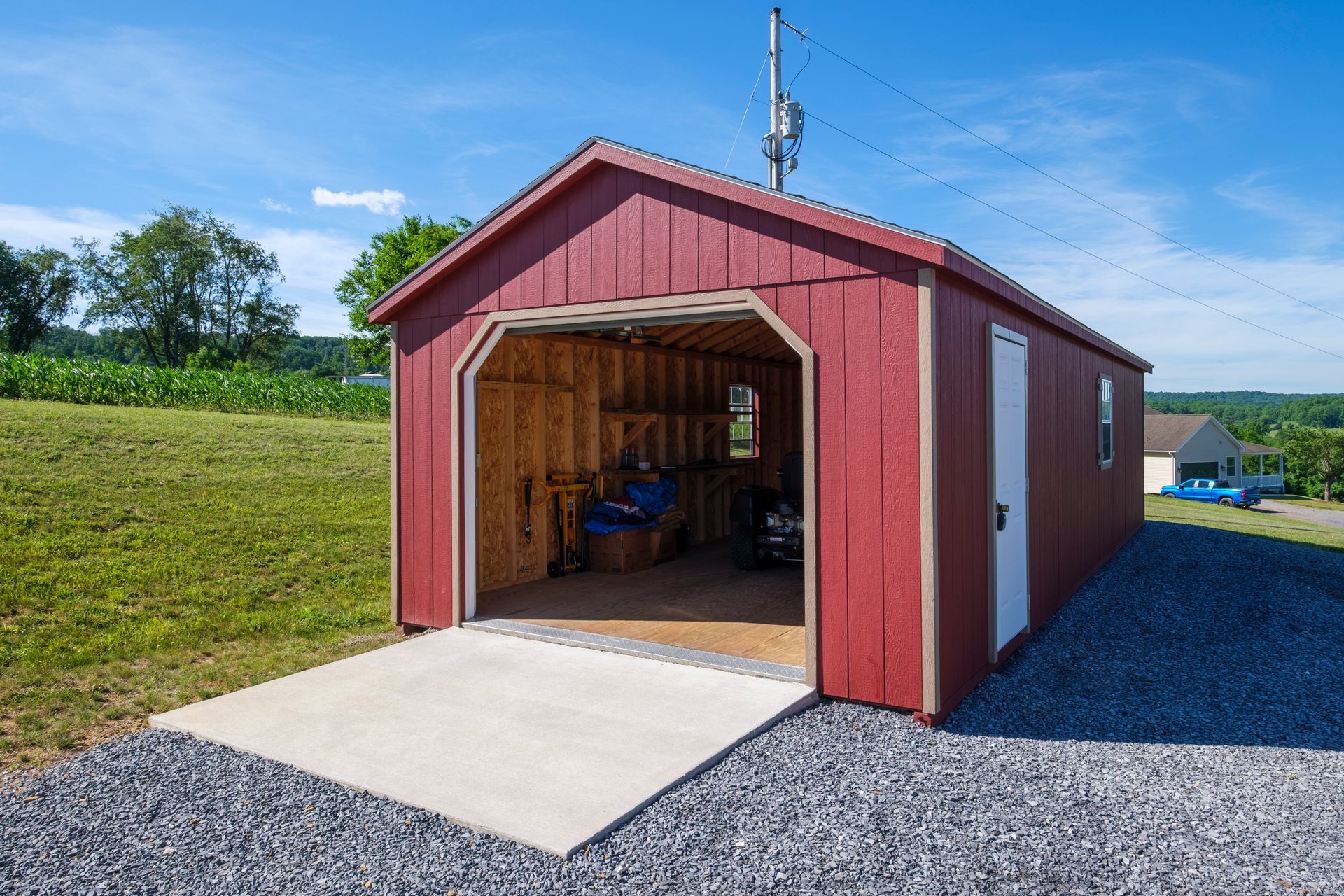 Red barn-style shed with open front, set on gravel beside a grassy slope under a blue sky