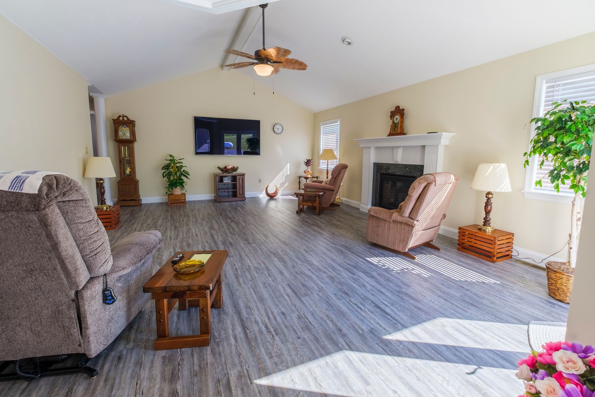 Spacious living room with gray sofa, fireplace, chairs, wall-mounted TV, and sunlight on wood floors