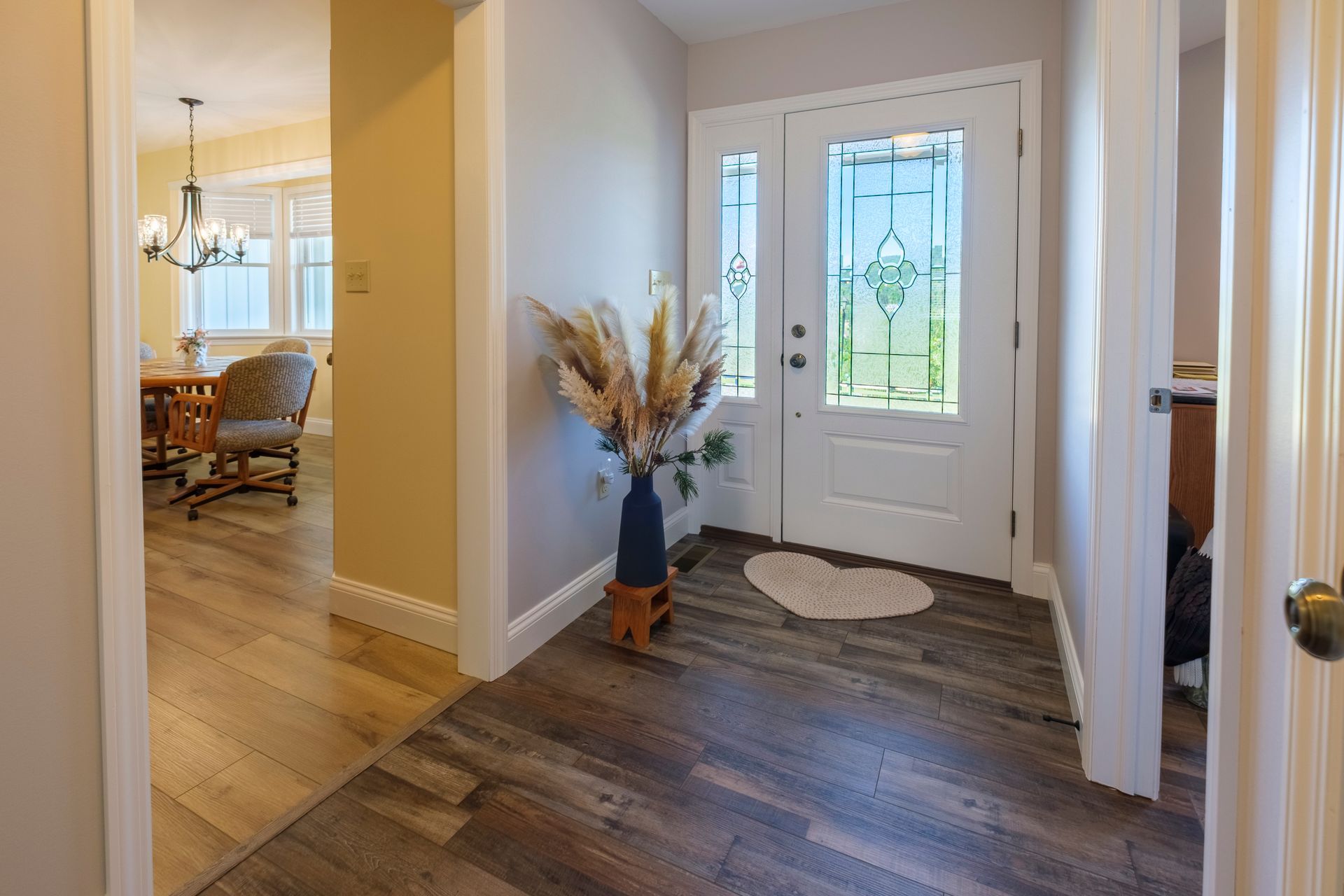 Front entryway with white door, dark wood floor, vase of dried flowers, and dining room visible nearby