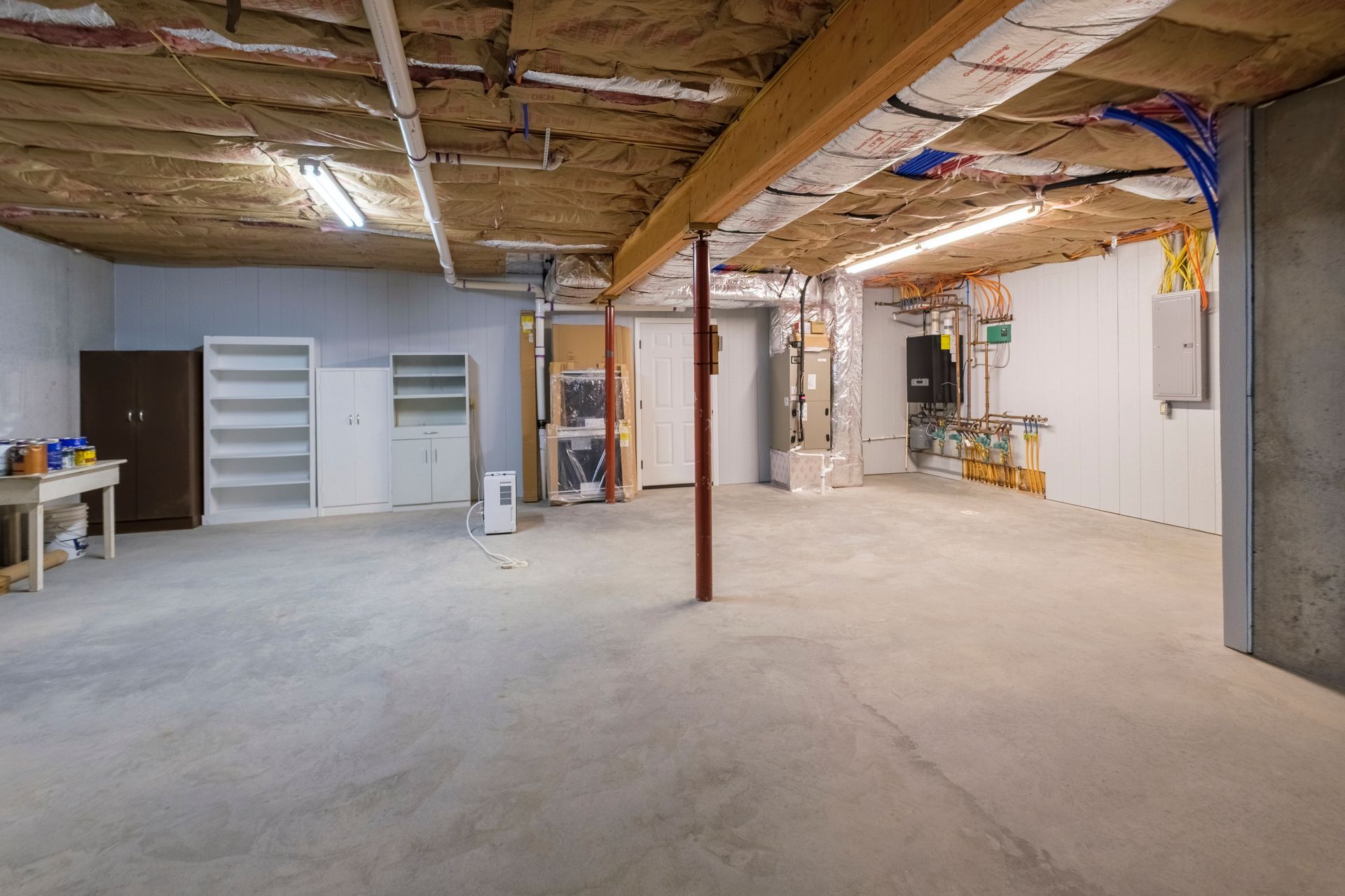 Empty unfinished basement with exposed beams, concrete floor, shelves, and utility posts